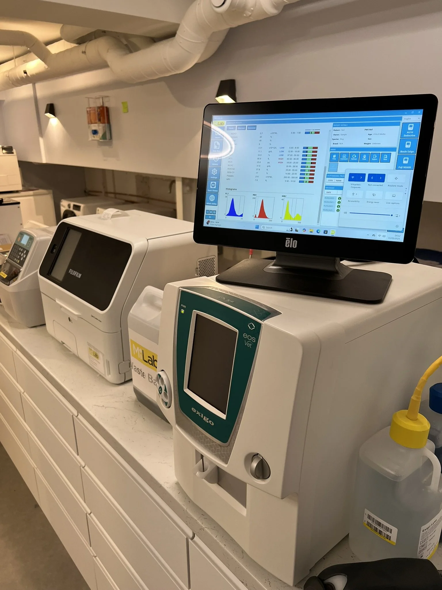 Laboratory equipment including an open medical analyzer with a monitor displaying test results, a blood or sample processor, and other lab devices on a white counter in a clinical setting.