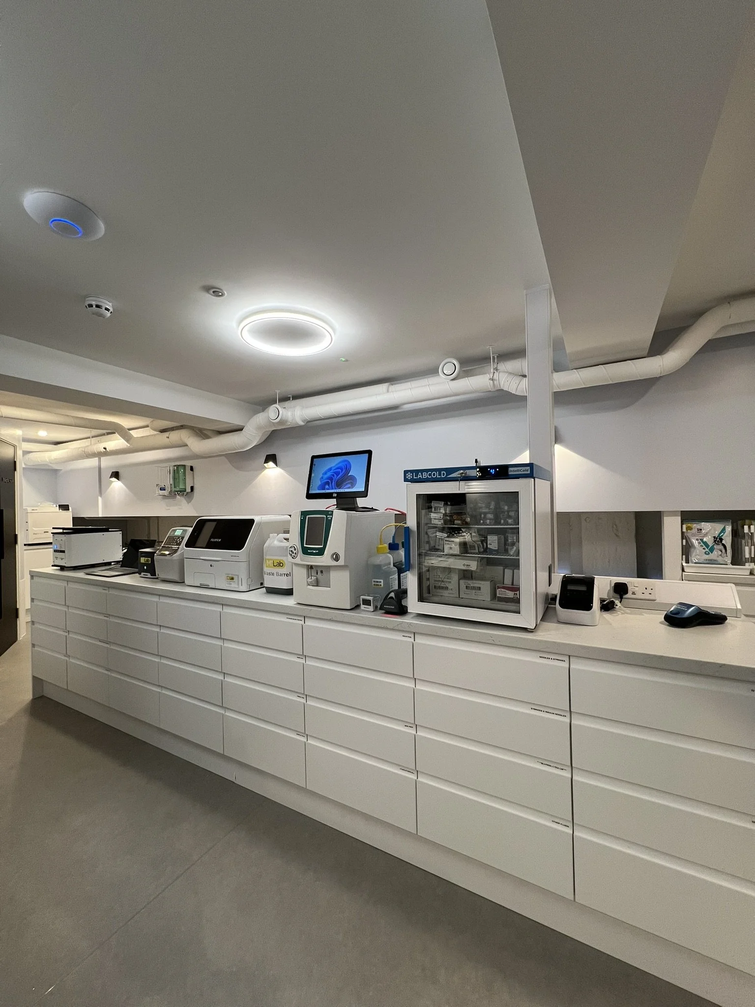 Hospital or laboratory work area with white counter, various medical and laboratory equipment, a computer monitor, and charts or supplies in a clean, modern setting.