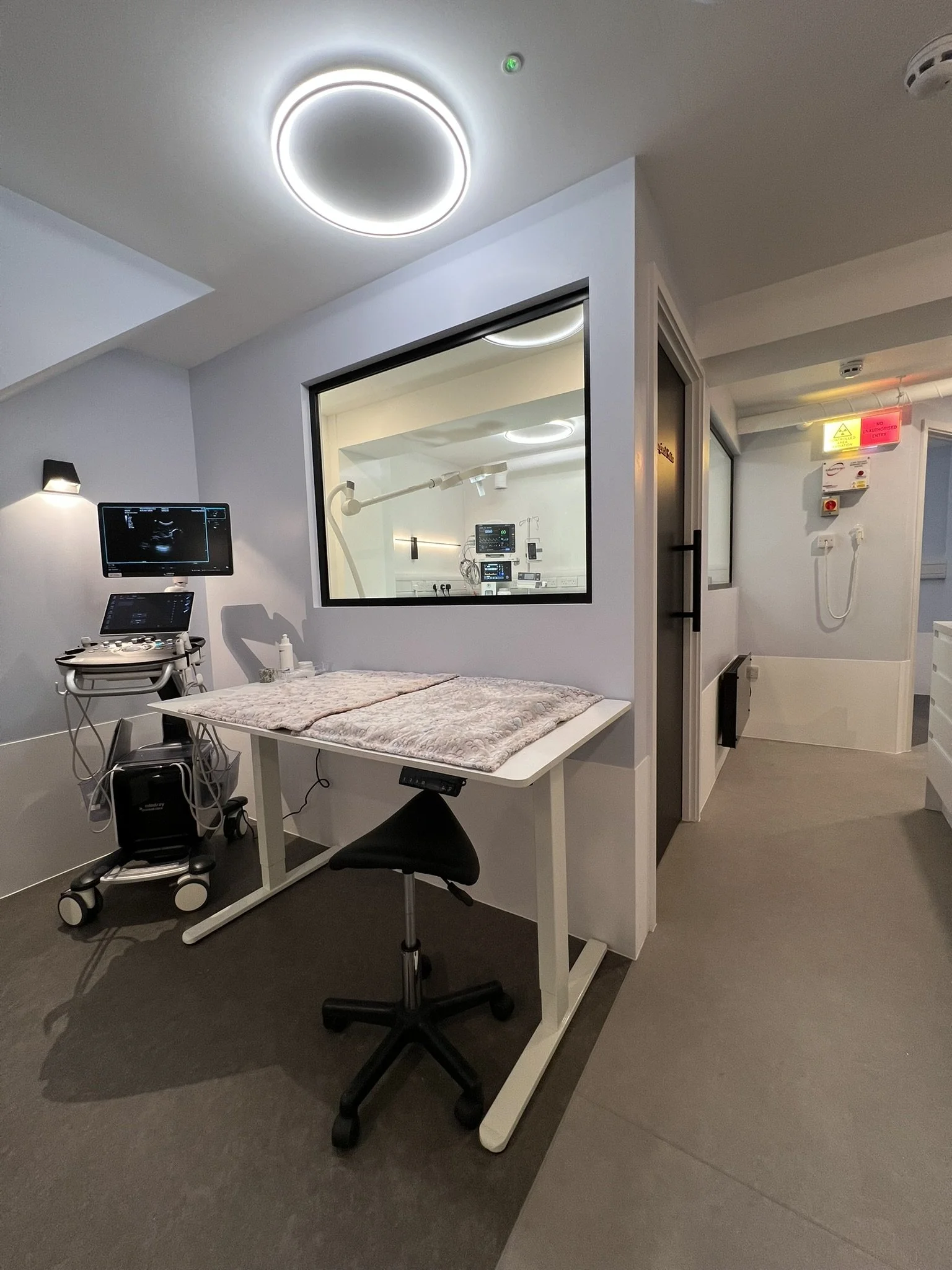 Medical examination room with ultrasound machine, examination table, and observation window to an adjacent room with medical equipment.