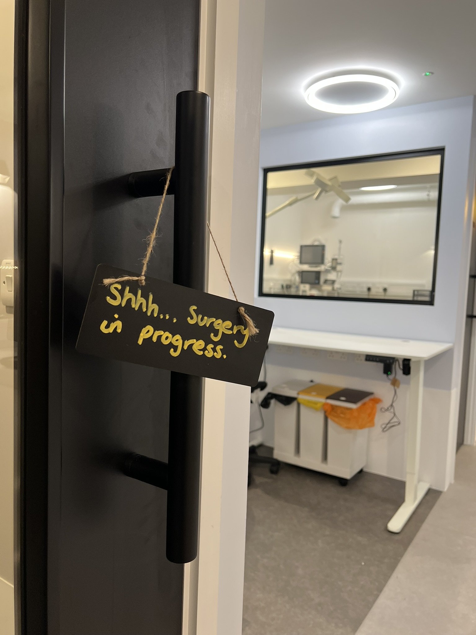 Hospital room with a sign on the door handle that reads 'Shhh... Surgery in progress' in yellow handwriting. Inside the room, there is a white desk with a black chair, a yellow and black folder, orange trash bag, and some electronics. A large window 