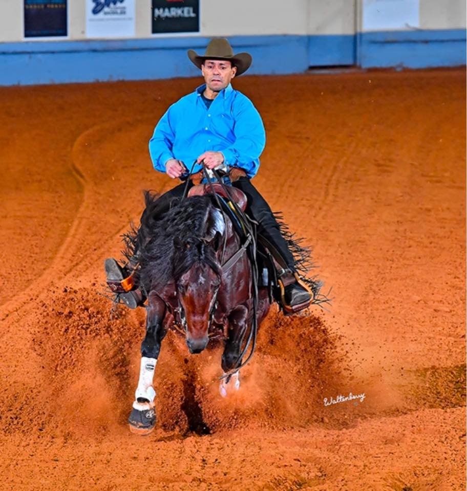 A man wearing a cowboy hat and blue shirt riding a galloping horse on a dirt indoor arena.