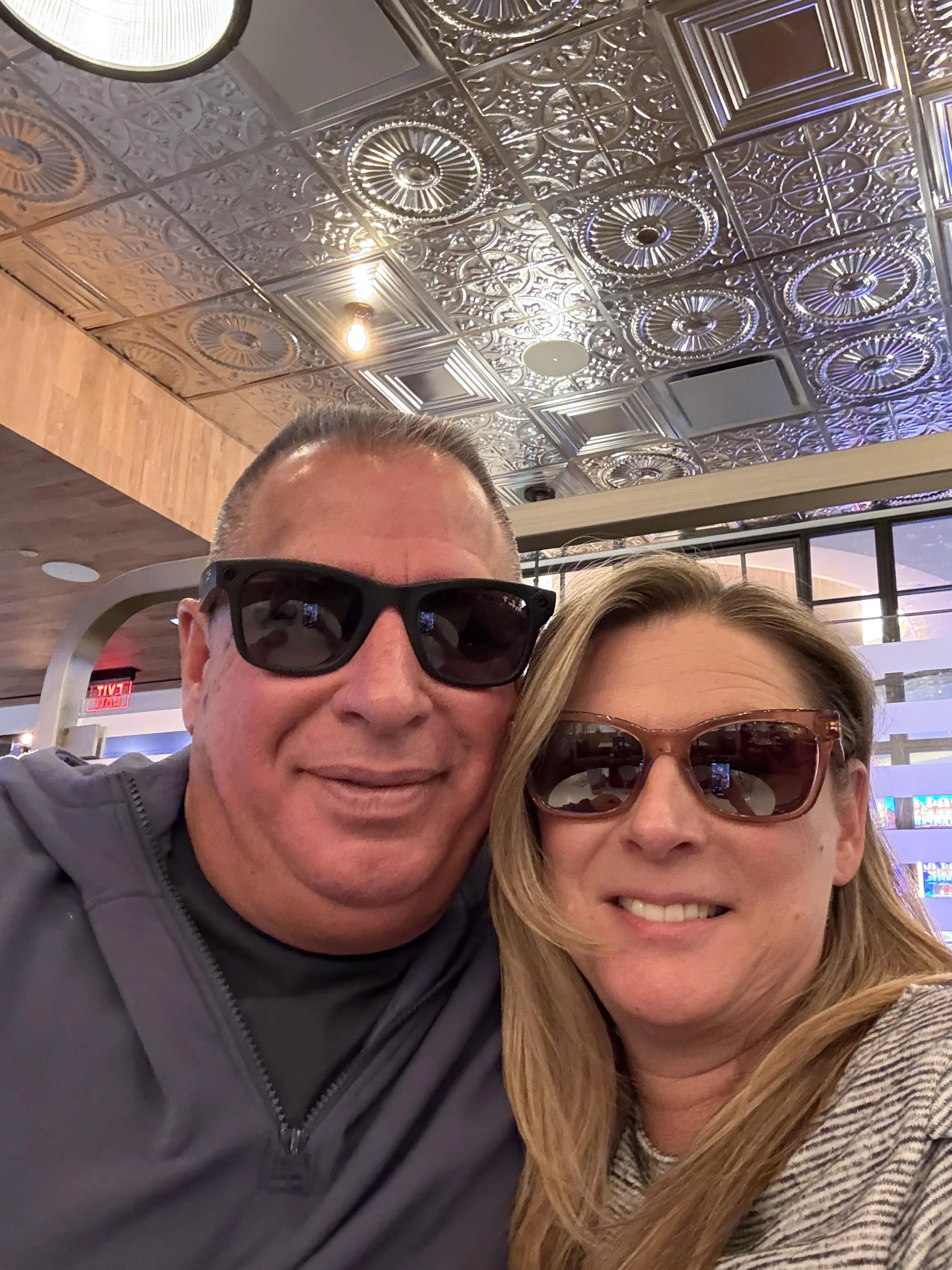 Close-up selfie of a man and woman wearing sunglasses in a restaurant with intricate metallic ceiling tiles.