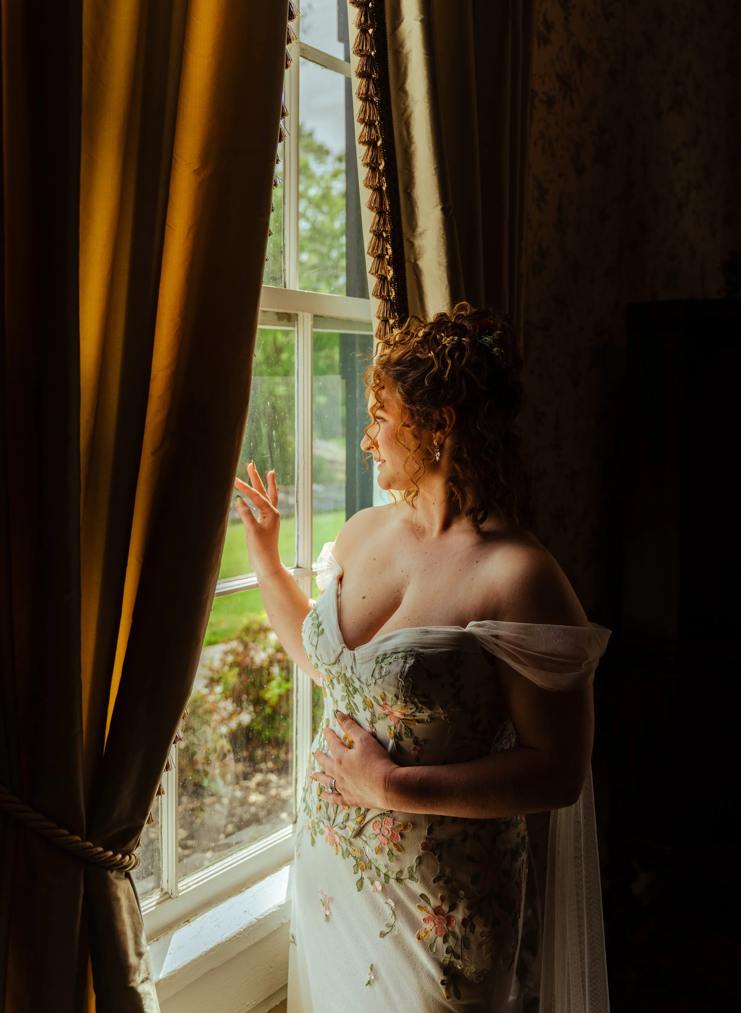 A woman with curly hair, wearing a dress with floral embroidery, stands by a window with heavy curtains, gazing outside with a gentle smile.