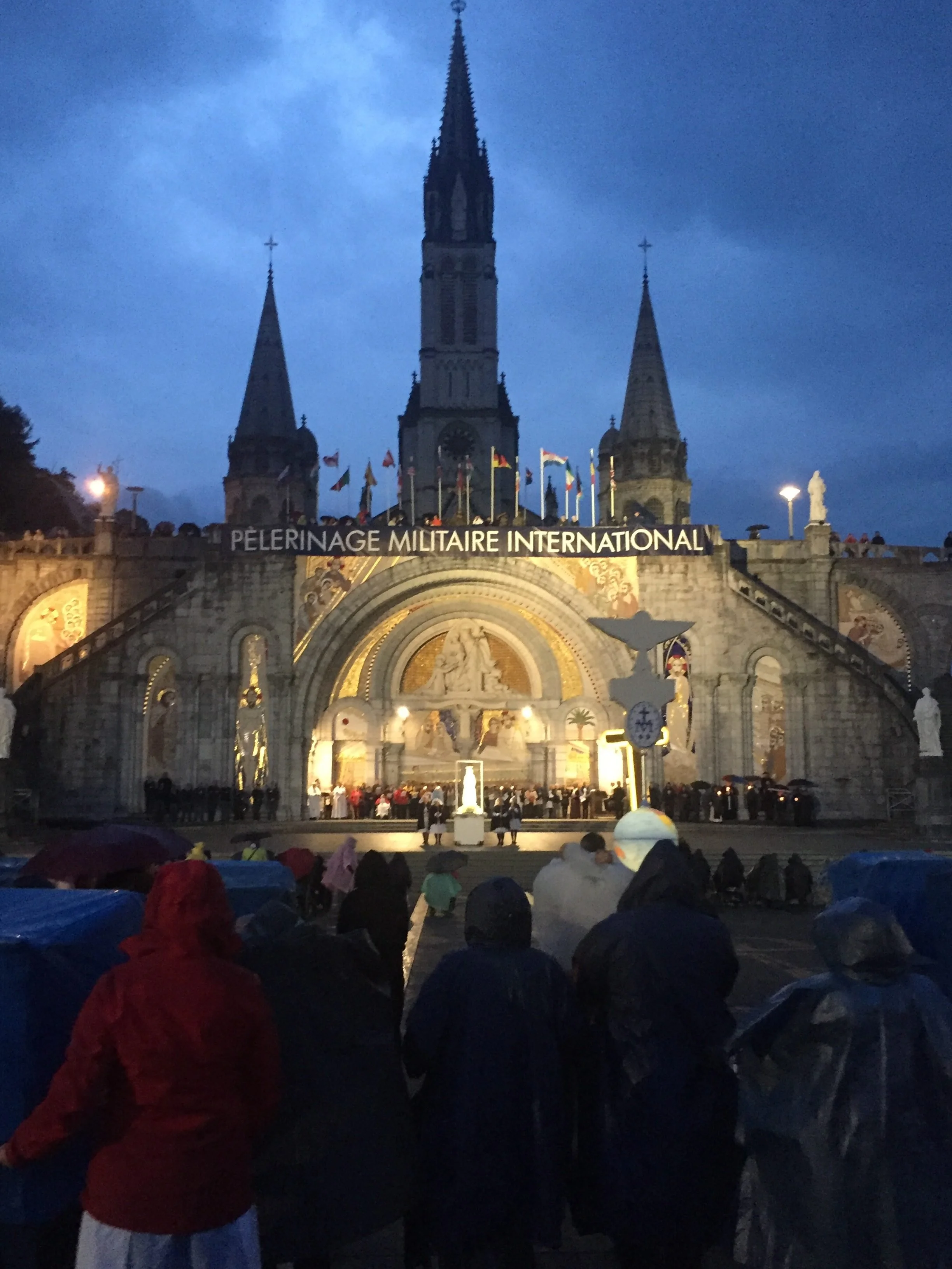 People gathered at the Basilica of Our Lady of the Rosary of Altötting, with a sign reading "Peleringue Militaire International," during an evening event with darkening skies.