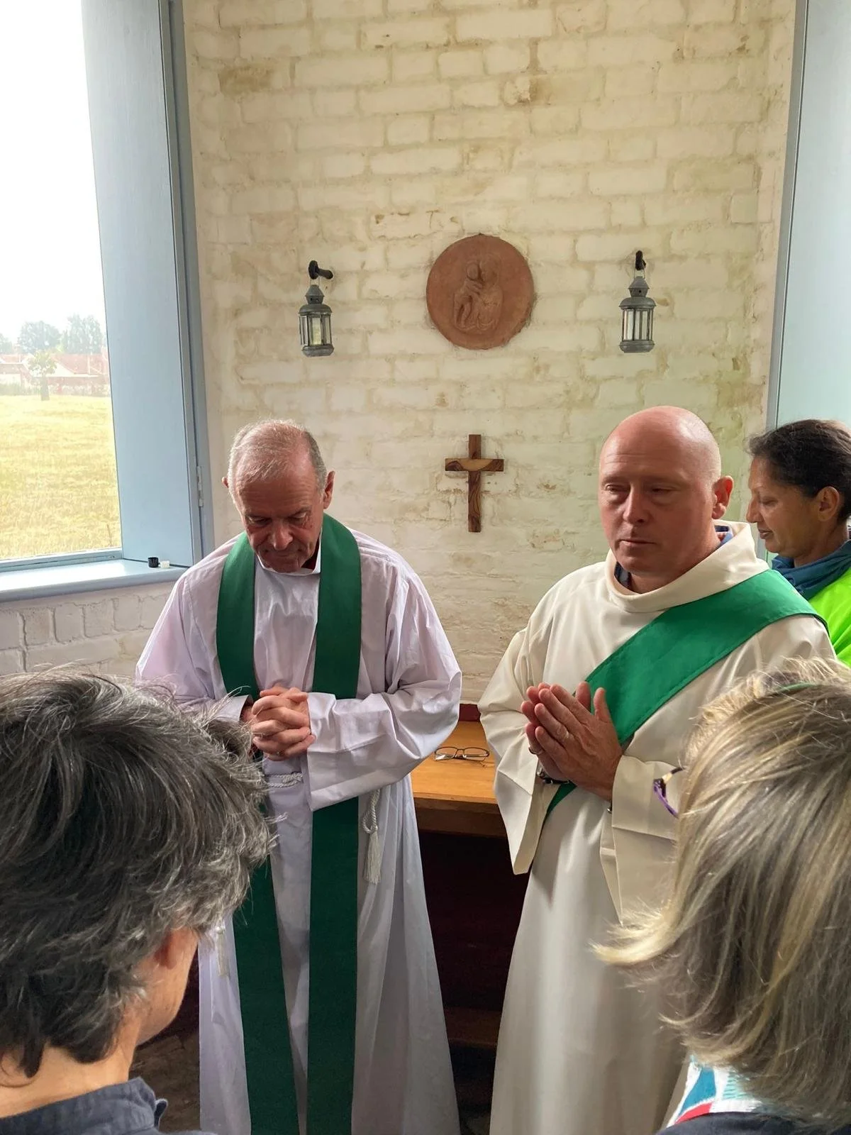 Two priests wearing white robes with green stoles and a woman in a bright green vestment kneel in prayer inside a small church or chapel. The background features a brick wall with a wooden cross, a religious plaque, and lantern wall sconces. A window reveals an outdoor field.