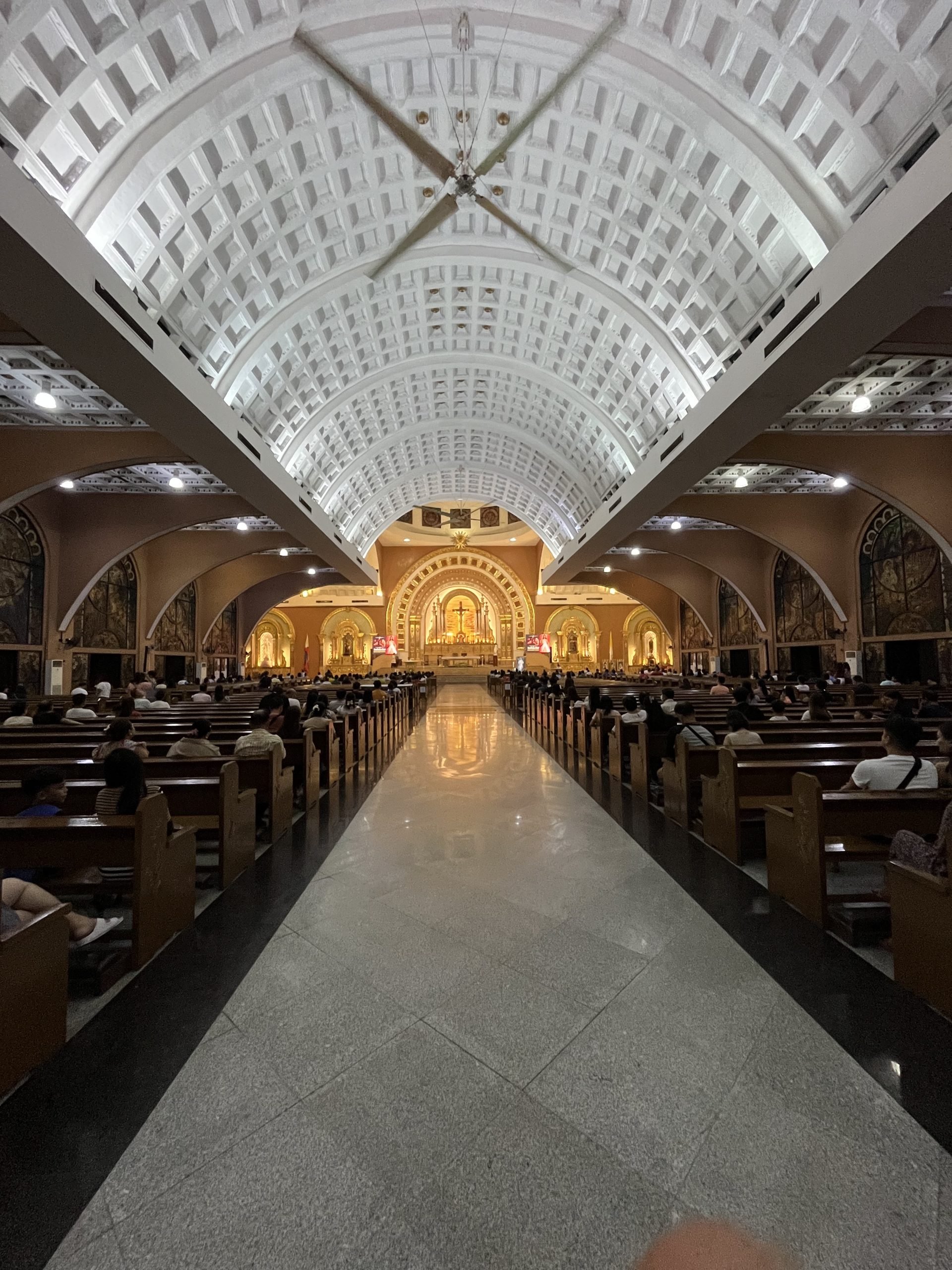 Interior of a church with a high vaulted ceiling, pews on either side, and an altar with religious imagery in the background.