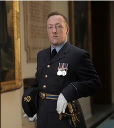 A man in a formal military uniform with medals standing indoors, posing with his hand on his hip.
