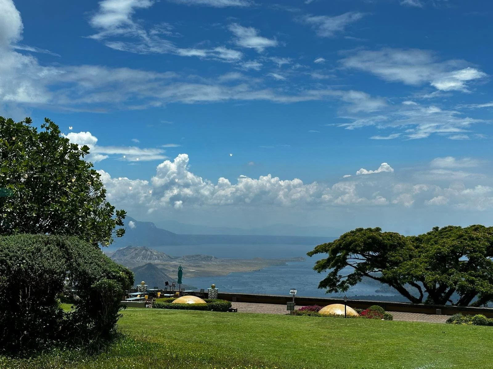 View of a scenic landscape with trees, green grass, a body of water, mountains in the background, and a partly cloudy sky.