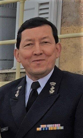 A man in a formal military uniform with ribbons and insignia, standing outdoors with a building and railing in the background.