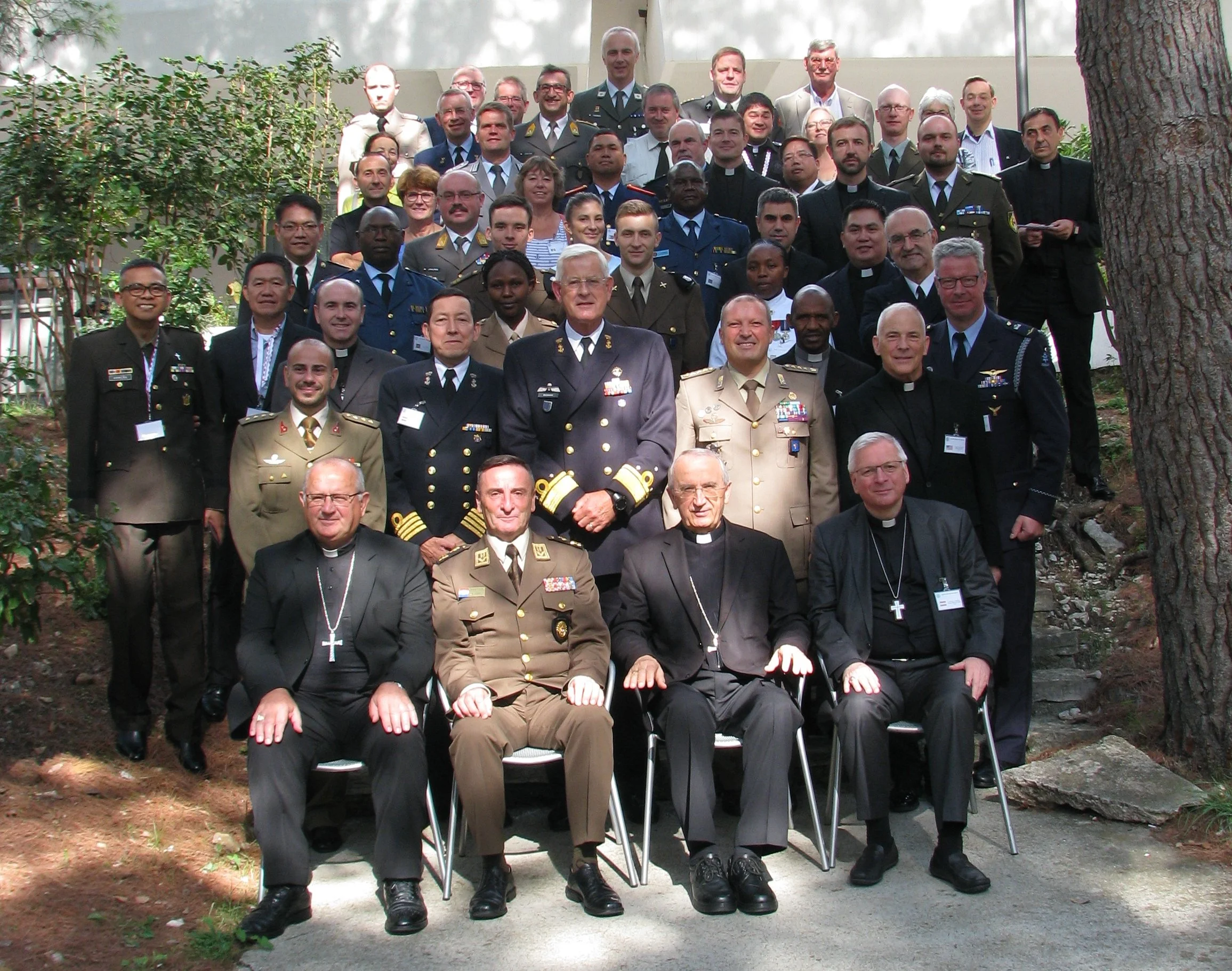 Group photo of military and religious leaders outdoors, with some seated and others standing, surrounded by trees and plants.