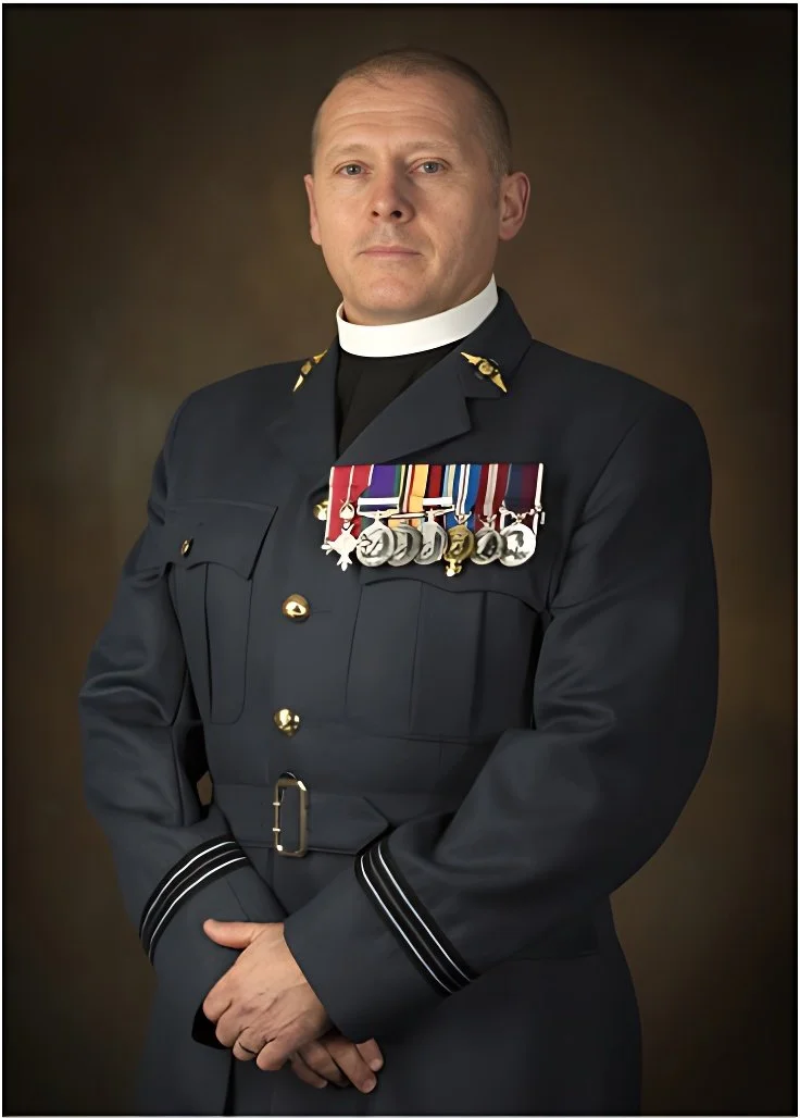 A man in a formal military uniform with medals and a white collar, standing with hands clasped in front of him, against a plain brown background.