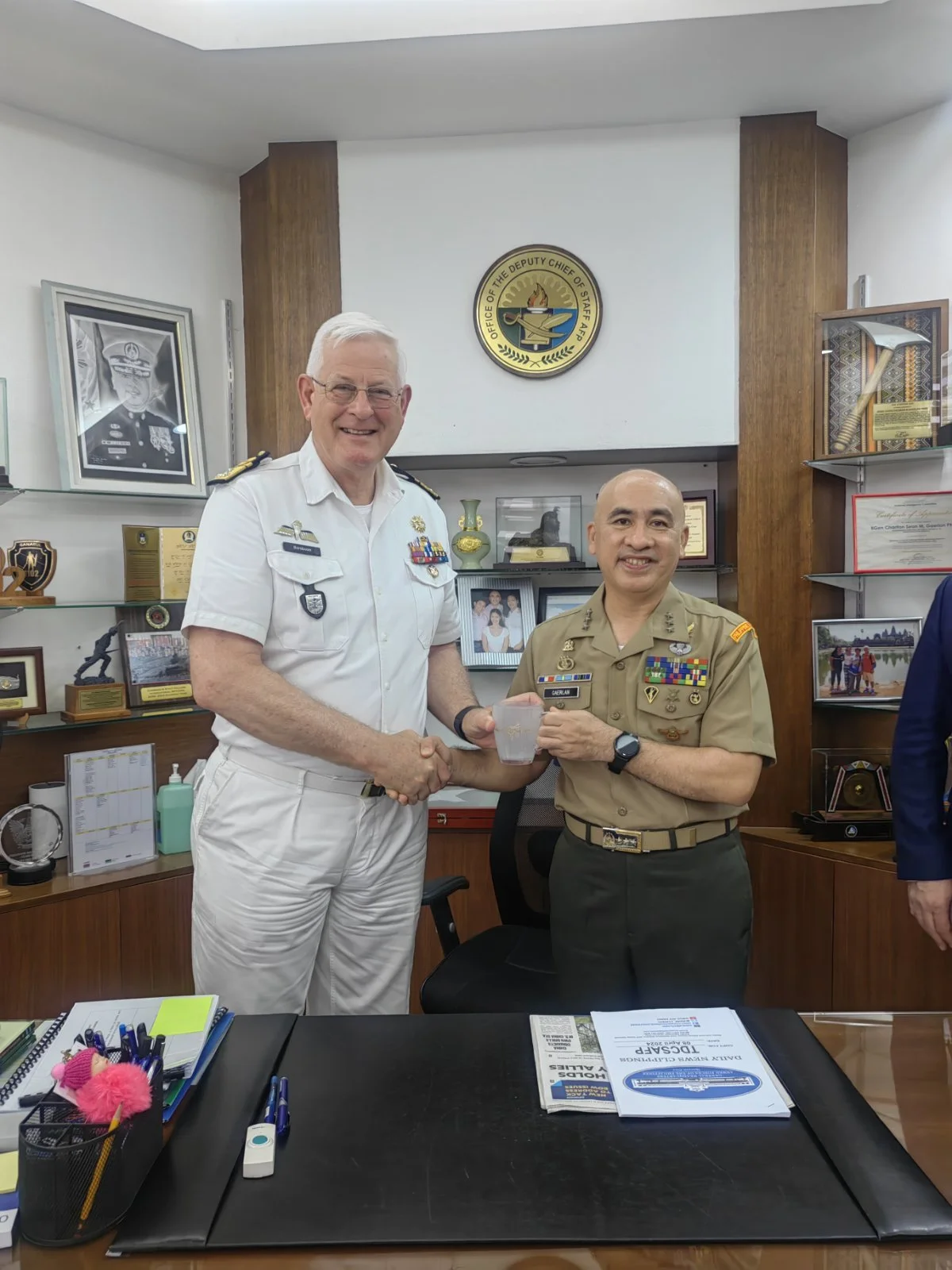 Two military officers shaking hands in an office, with the officer on the left holding a clear cup. The room has framed photographs, awards, and awards displays on the shelves behind them.