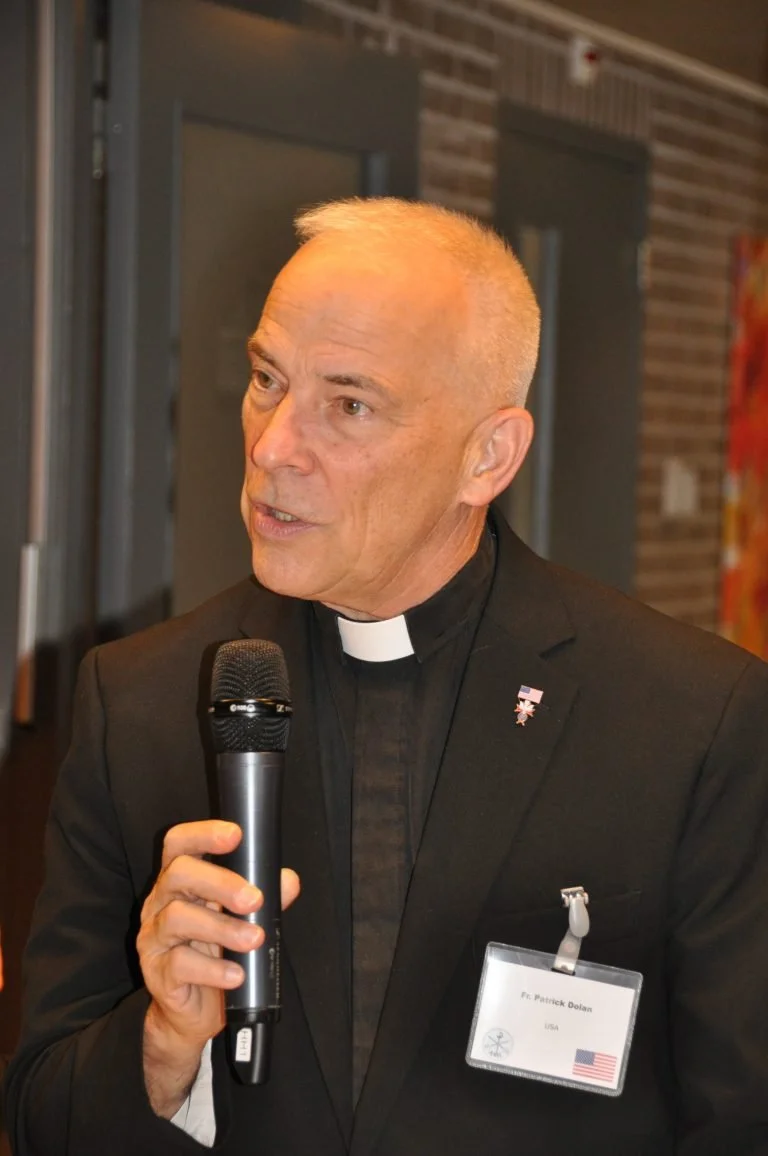 A clergyman, wearing a black suit with a clerical collar, is speaking into a handheld microphone at an indoor event. He has short, gray hair and wears a name badge and a pin featuring the American flag.