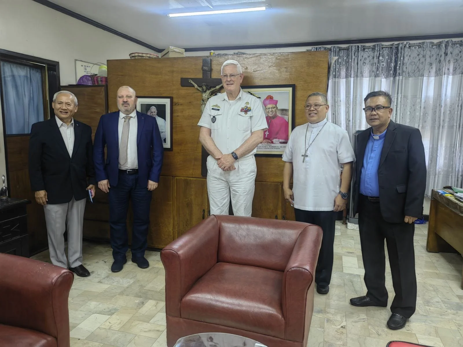 Group of six men in formal and religious attire standing in an office with framed photographs, a crucifix, curtains, and a wooden wall behind them.