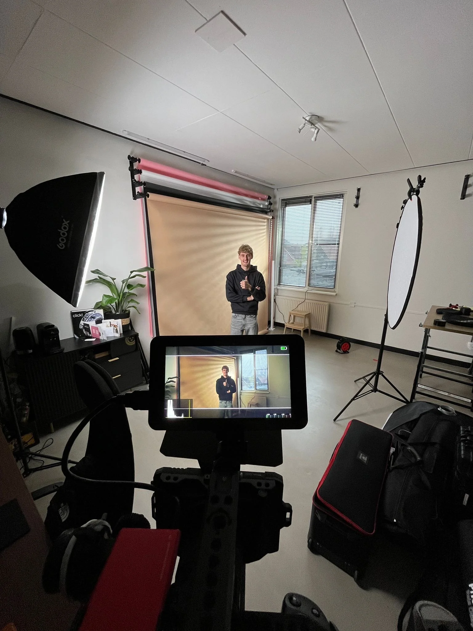 Cameraplek met een camera op een statief die een jongen in een fotoshootruimte filmt of fotografeert, met een scherm dat het beeld laat zien van de jongen voor een beige achtergrond. Er is foto- of filmverlichting en een grote paraplu-licht, een raam met jaloezieën en verschillende apparatuur.