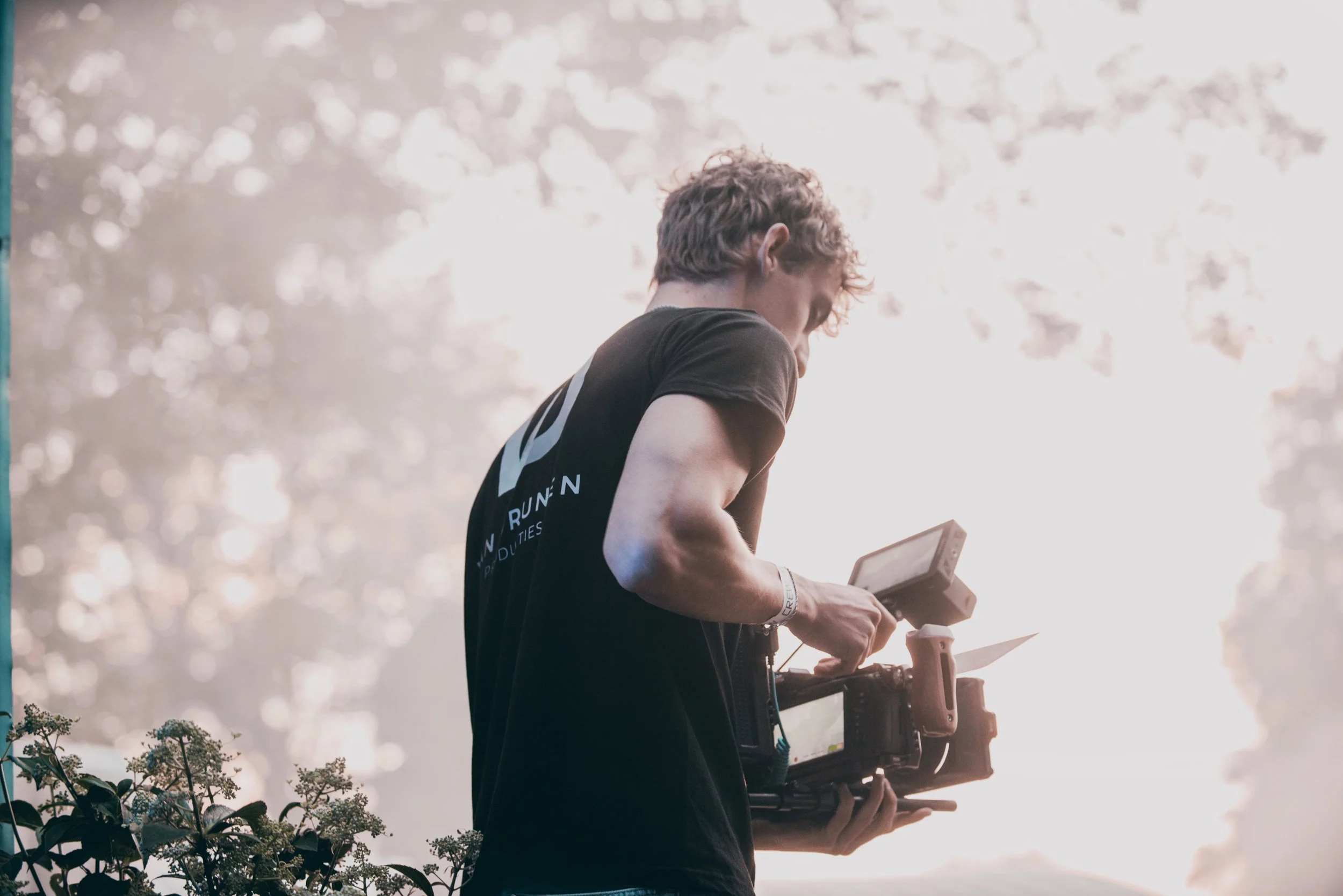 Een man houdt een filmcamera vast en lijkt te filmen of te fotograferen, met een lichte achtergrond en bomen op de achtergrond.