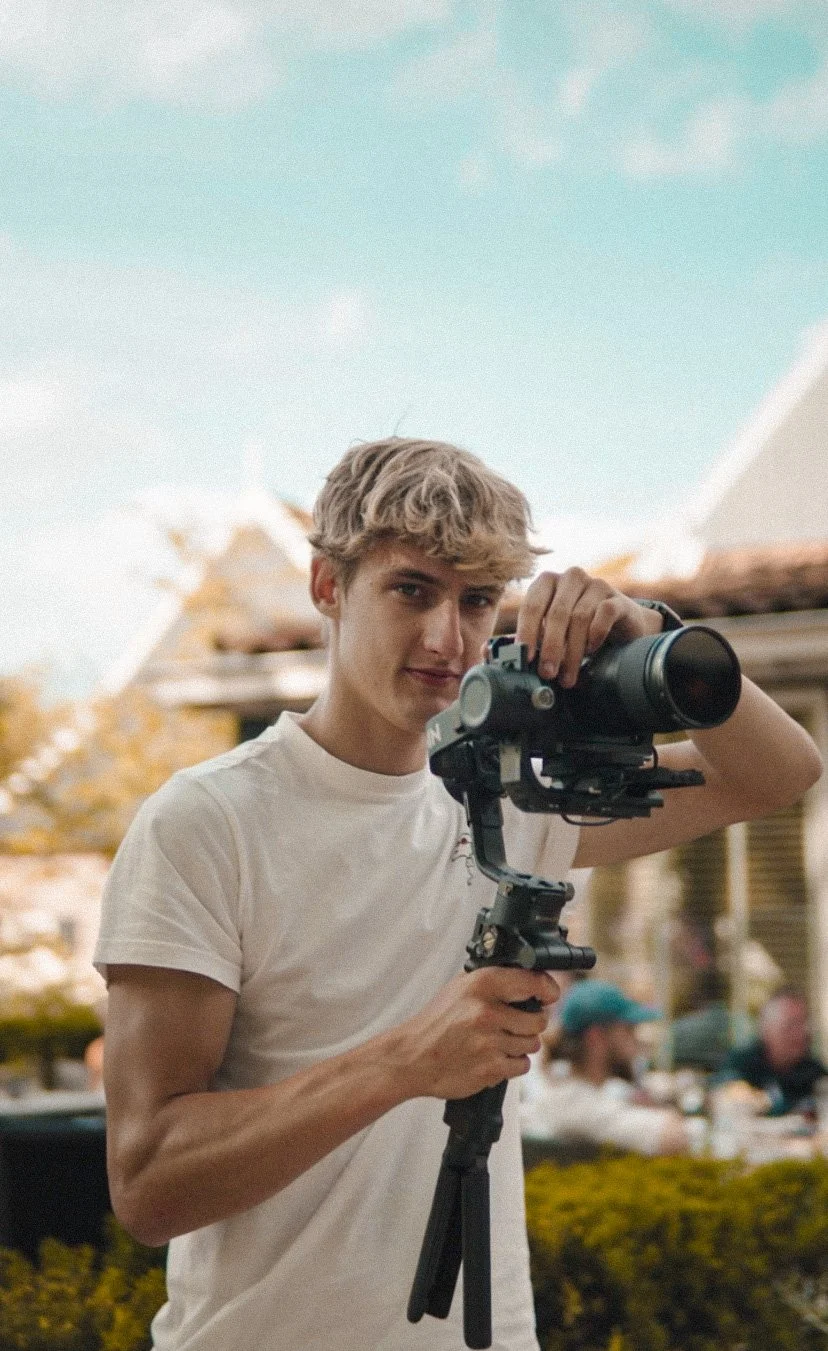 Jongen met blond krullend haar houdt een camera op een statief en kijkt door de lens.