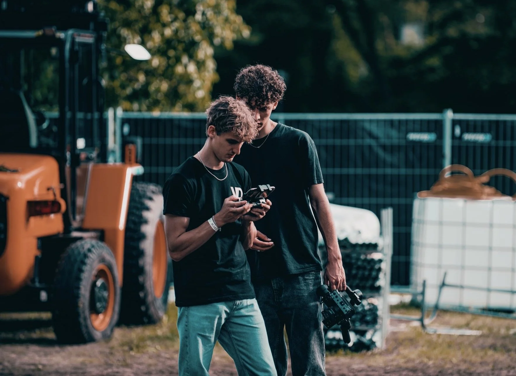 Twee jonge mannen kijken naar een smartphone en een camera in een buitenomgeving met een hek en een grote machine op de achtergrond.