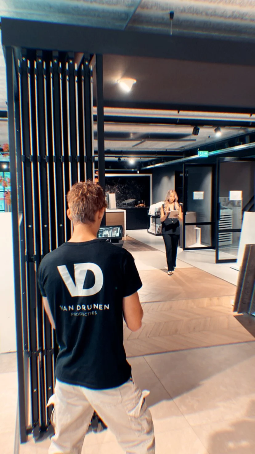 Persoon met 'Van Drunen' T-shirt filmt een vrouw met tablet in interieur van een modern zaakcentrum.