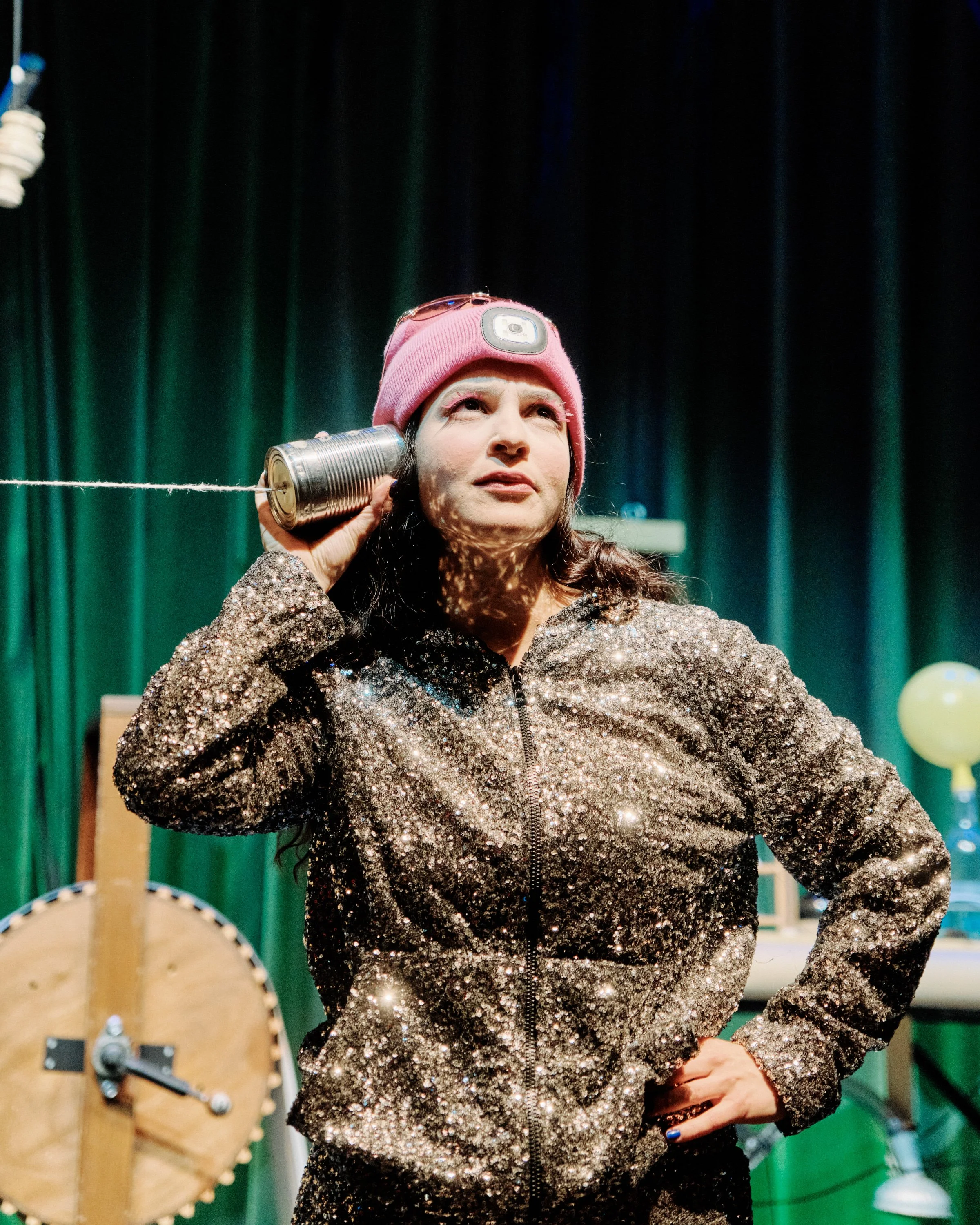 Frau mit pinker Mütze und Glitzerjacke hält eine Dose an das Ohr auf einer Bühne mit grünen Vorhängen.