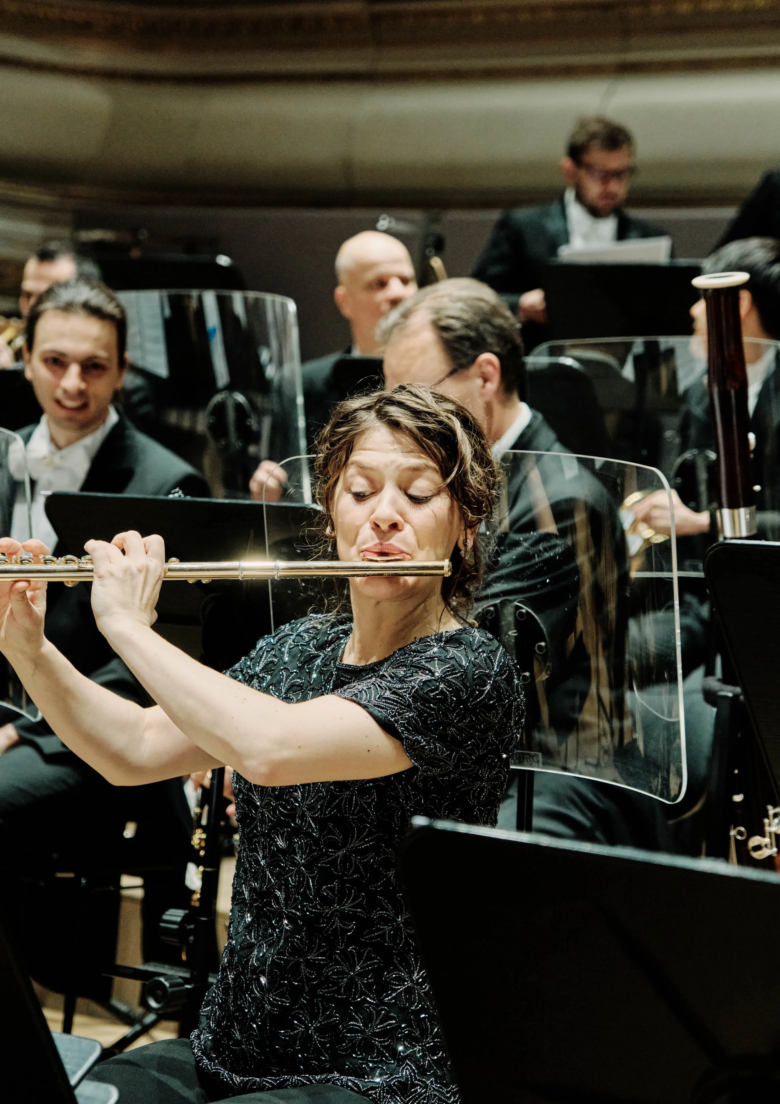 Eine Frau spielt Flöte in einem Orchester, umgeben von aufmerksamen Musikern in formeller Kleidung.