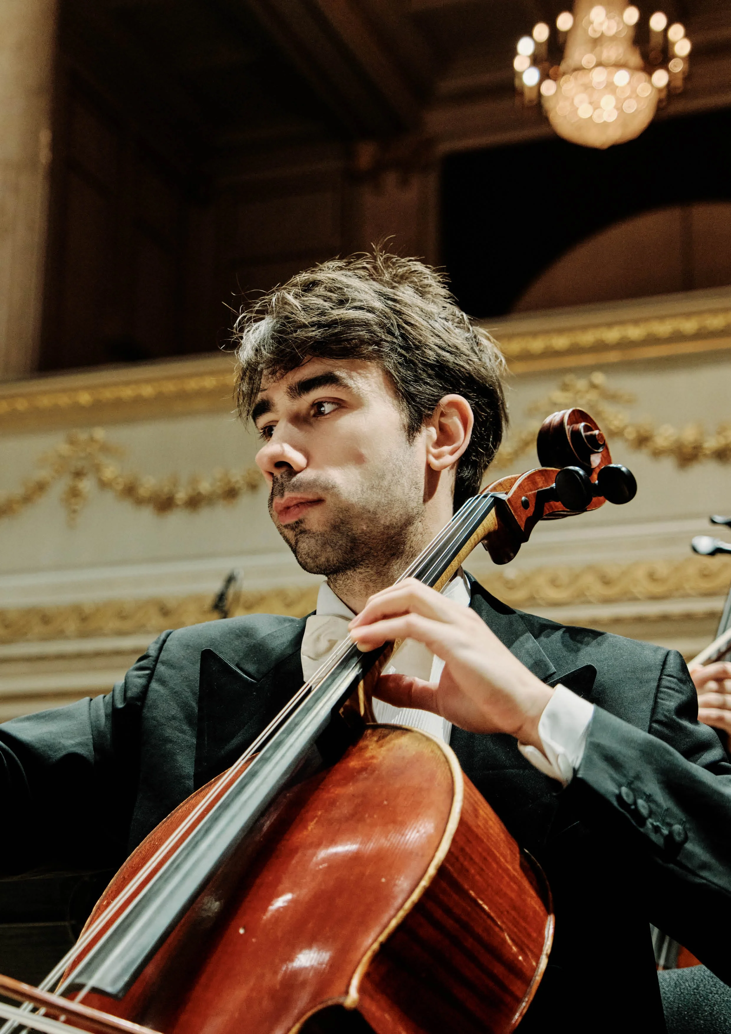Ein junger männlicher Cellist in formeller Kleidung spielt bei einem Konzert in einem eleganten Saal mit goldverzierten Details.