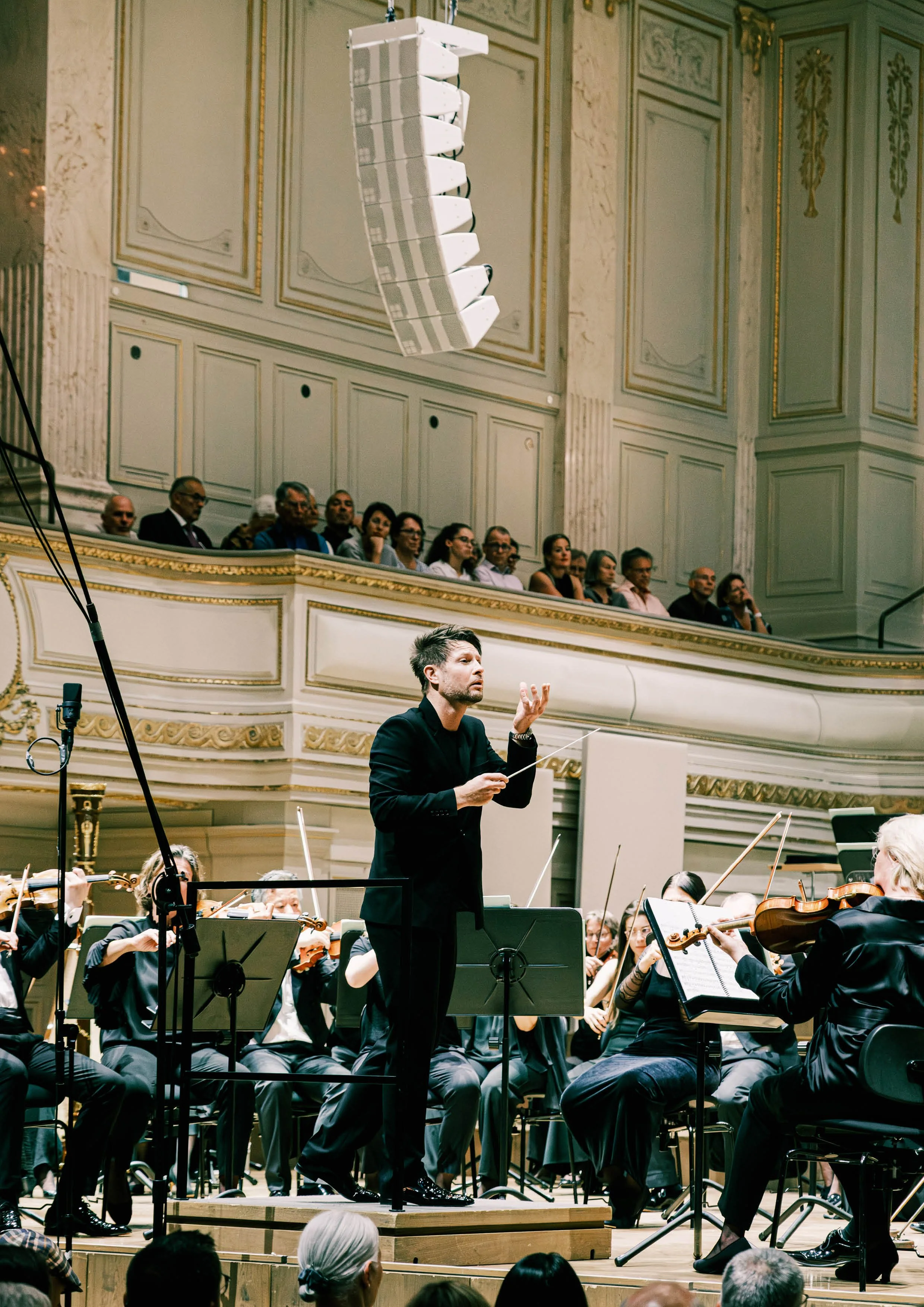 Ein Orchester in einem Konzertsaal, mit einem Dirigenten in schwarzer Kleidung, der die Musiker leitet. Es sitzen mehrere Musiker mit Violinen und anderen Instrumenten. Im Hintergrund sind Zuschauer im Balkon zu sehen.