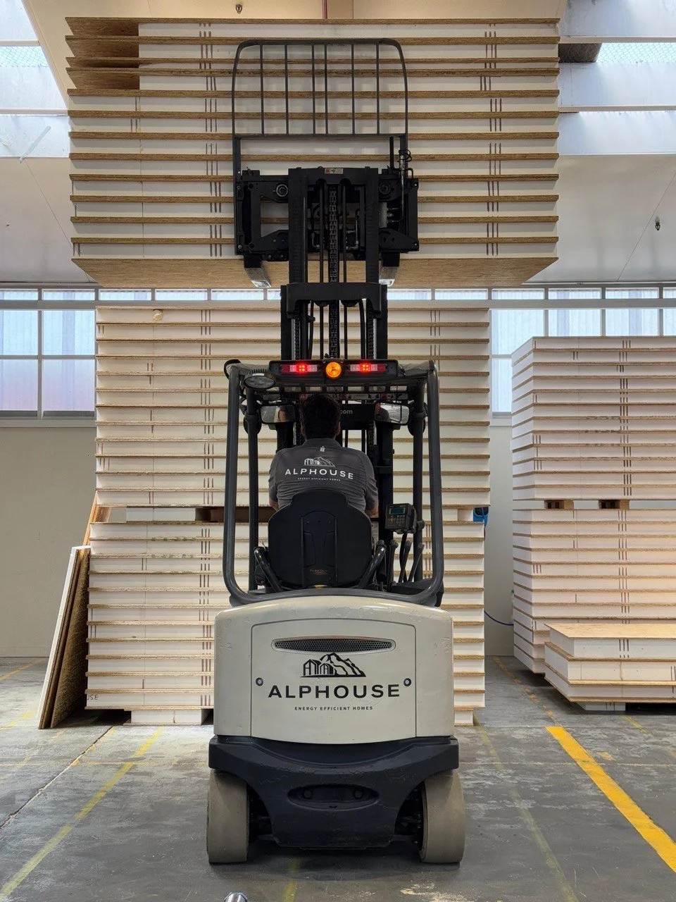 A forklift operator in an indoor warehouse moving stacks of wooden panels or boards.