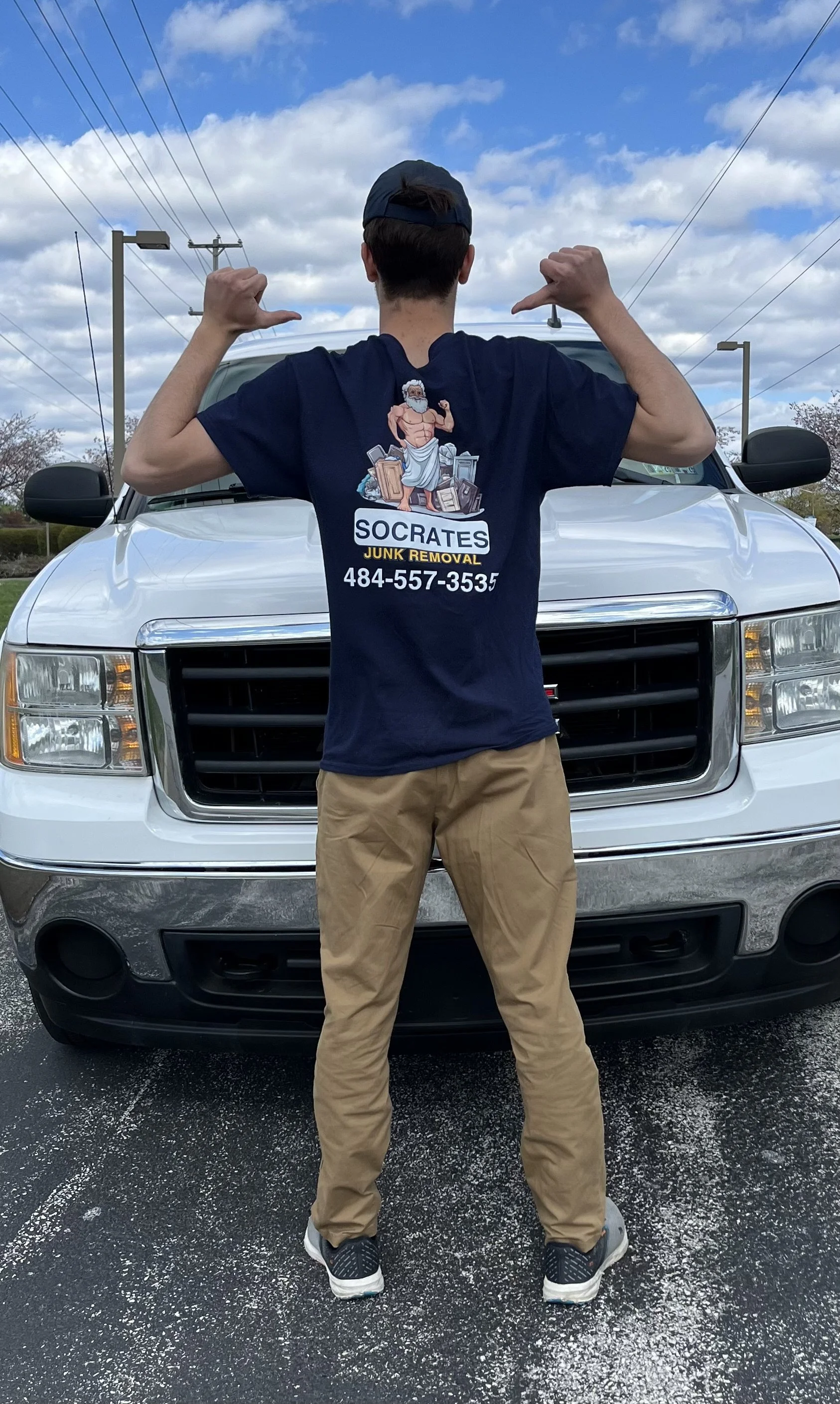 A person standing in front of a white pickup truck, flexing arms, wearing a navy blue t-shirt that says Socrates Junk Removal with a phone number, and a dark cap.