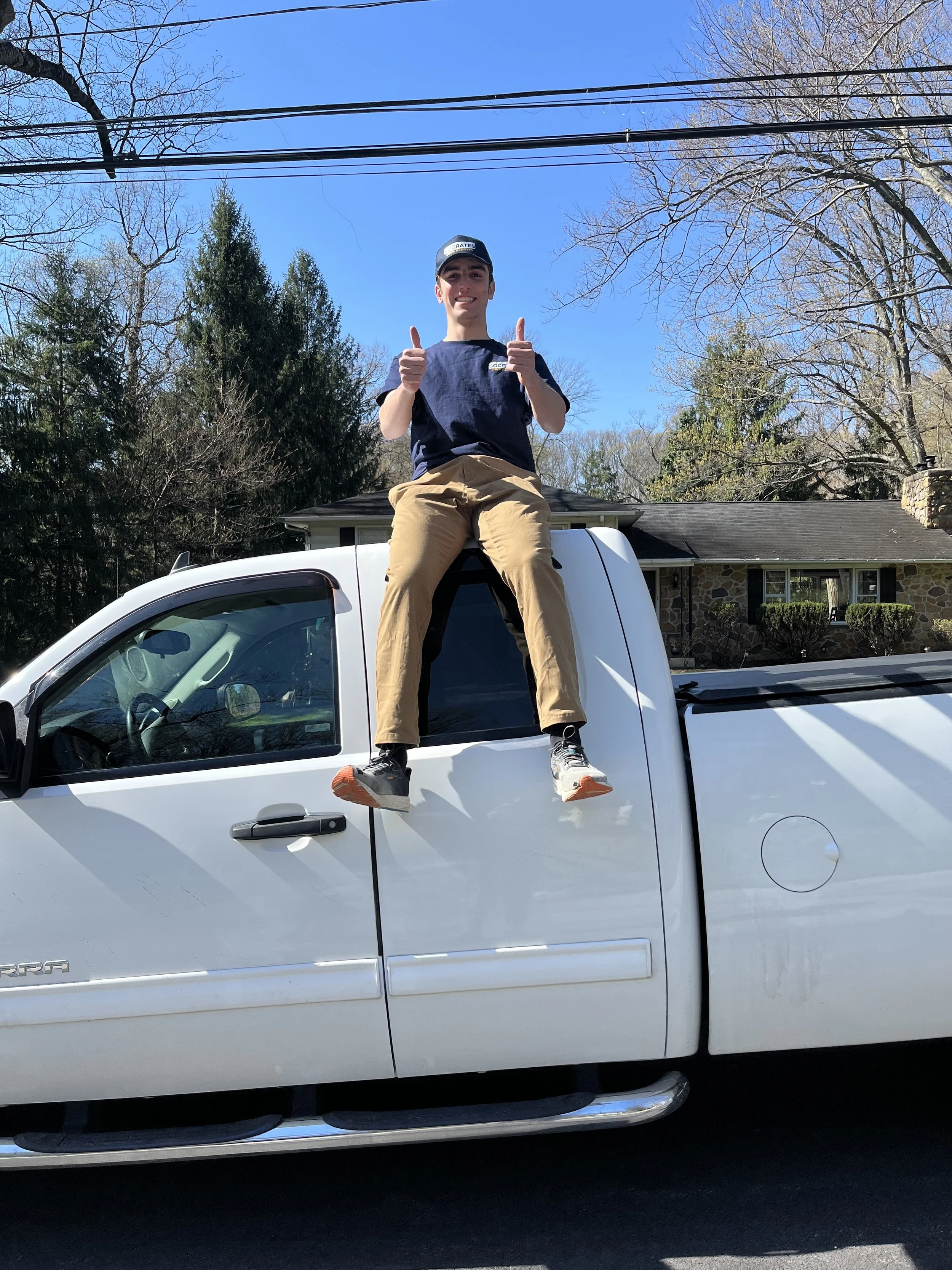 A young man is sitting on the roof of a white pickup truck, smiling and giving a thumbs-up with both hands, in a suburban neighborhood under clear blue skies.
