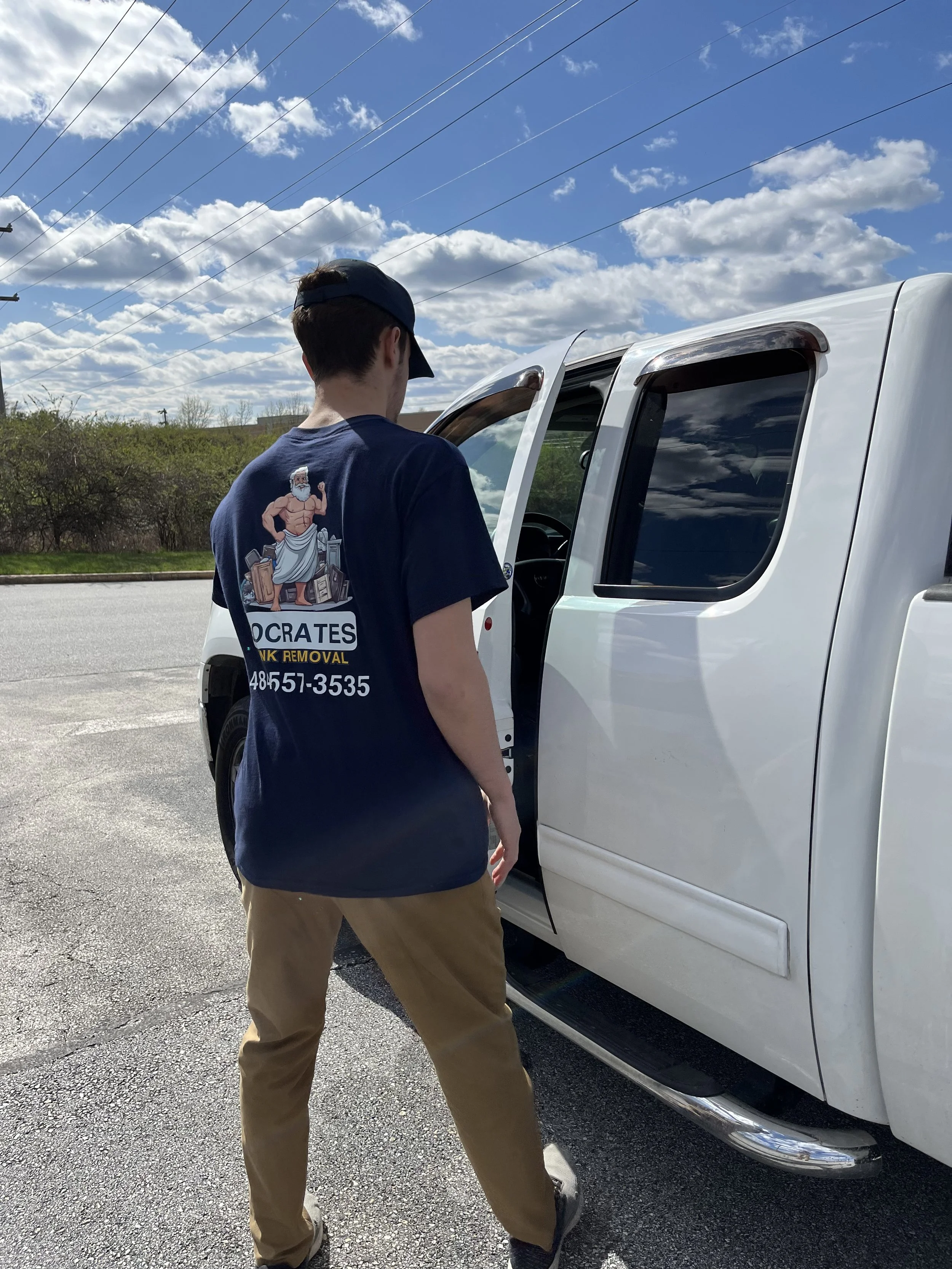 A person with a dark blue T-shirt and khaki pants opening the door of a white pickup truck. The back of the T-shirt has a graphic of a muscular man with a beard, wearing a toga, holding a hammer, with the words 'CROATES' and a phone number underneath.