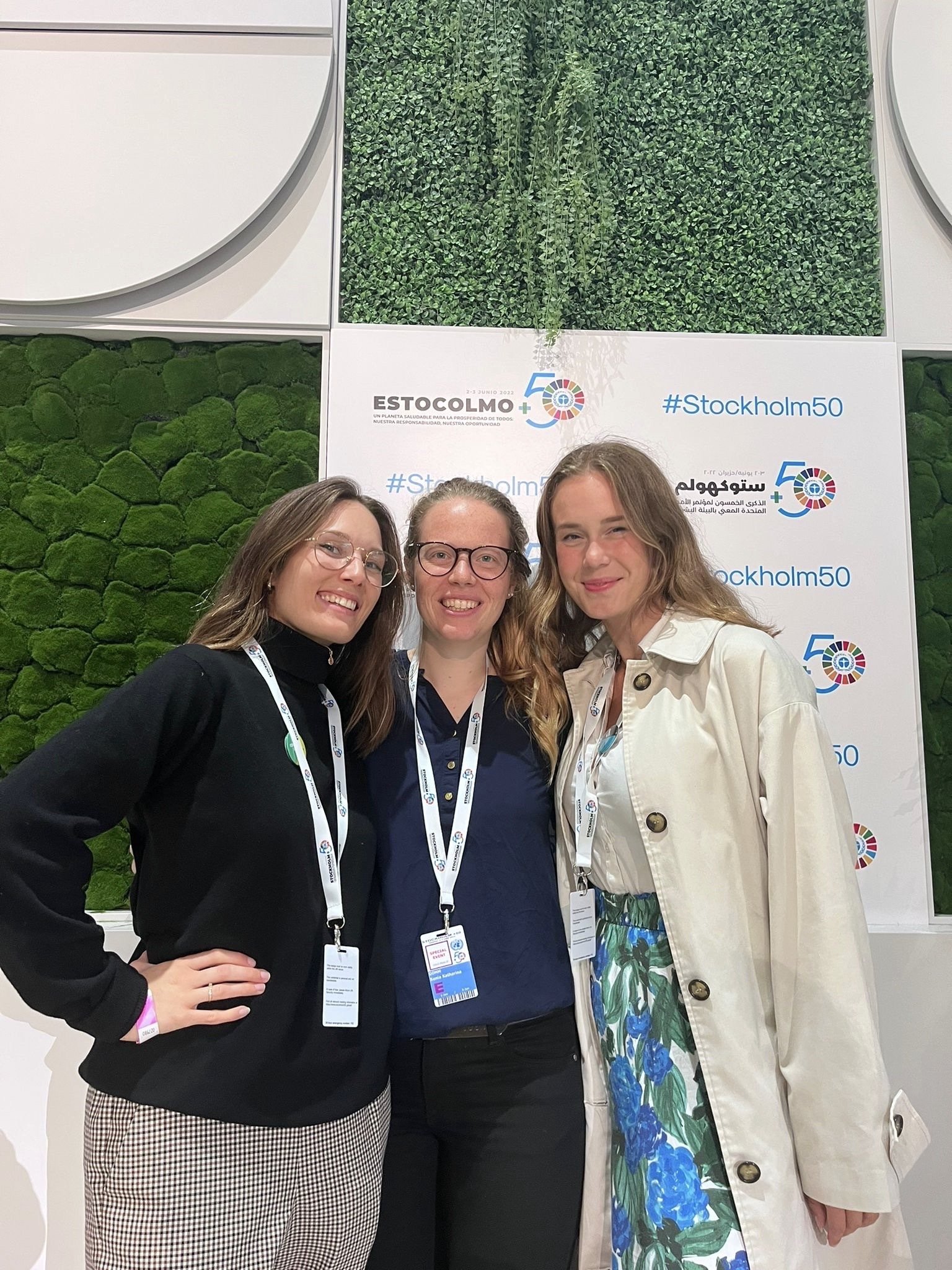 Tre personer står framför en vägg dekorerad med grönska och en skylt med texten "Estocolmo 50" och "#Stockholm50". De har namnbrickor runt halsen.