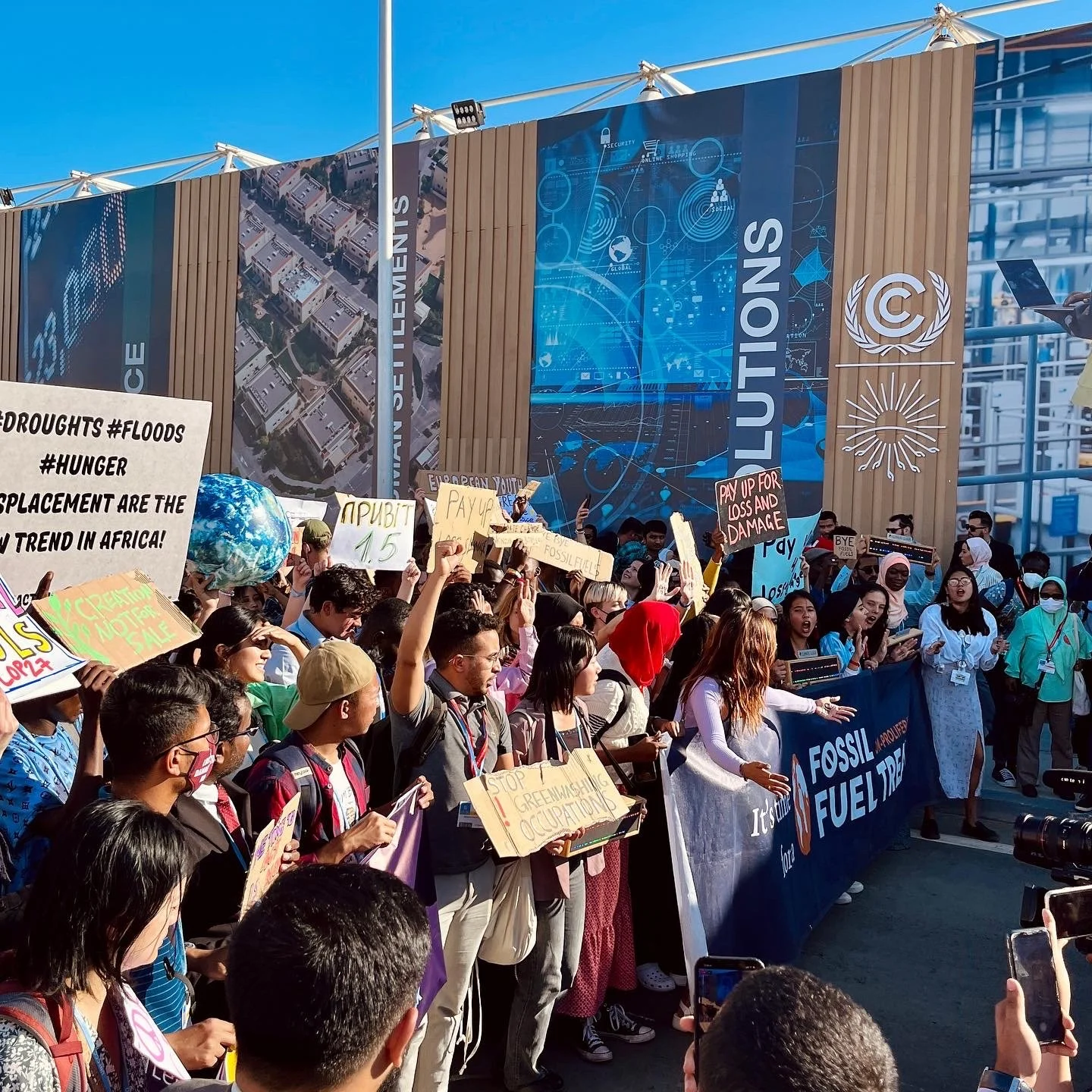 En grupp människor deltar i en protest, håller upp skyltar och banderoller med budskap mot klimatförändringar och fossilt bränsle, framför en byggnad med affischer om lösningar.