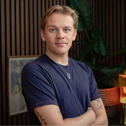 Person står med korslagda armar, klädd i en mörkblå t-shirt, framför en bakgrund med trämönstrade paneler och gröna växter.