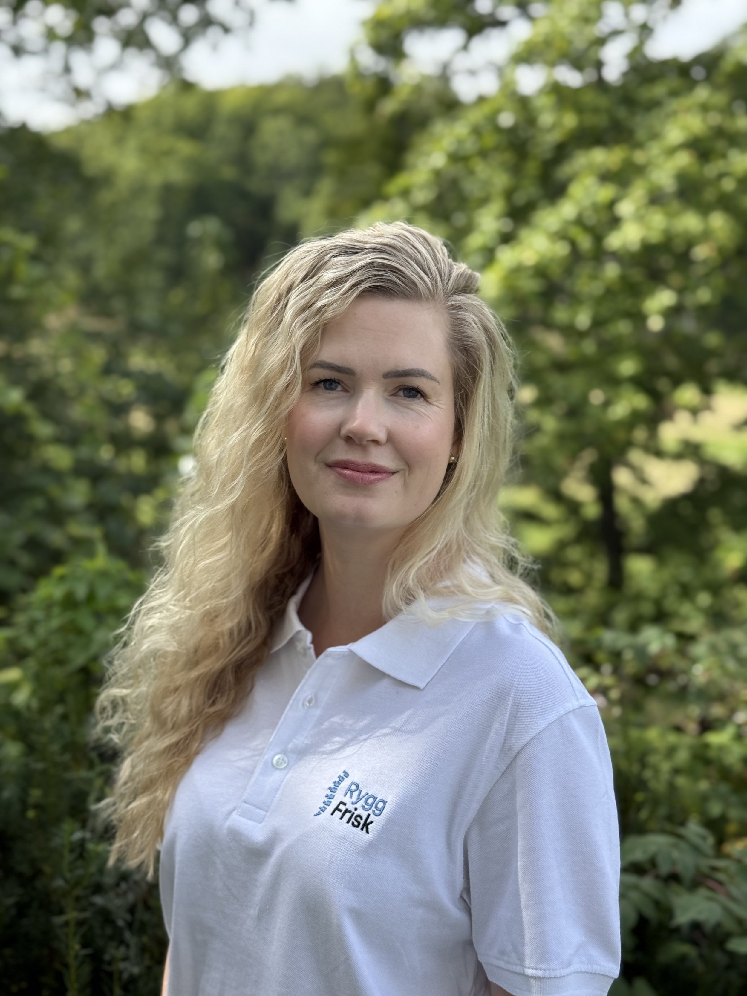 En kvinne med blonde hår smiler foran grønne trær, hun har på seg en hvit polo-trøye med tekst som sier "Rygg Frisk".