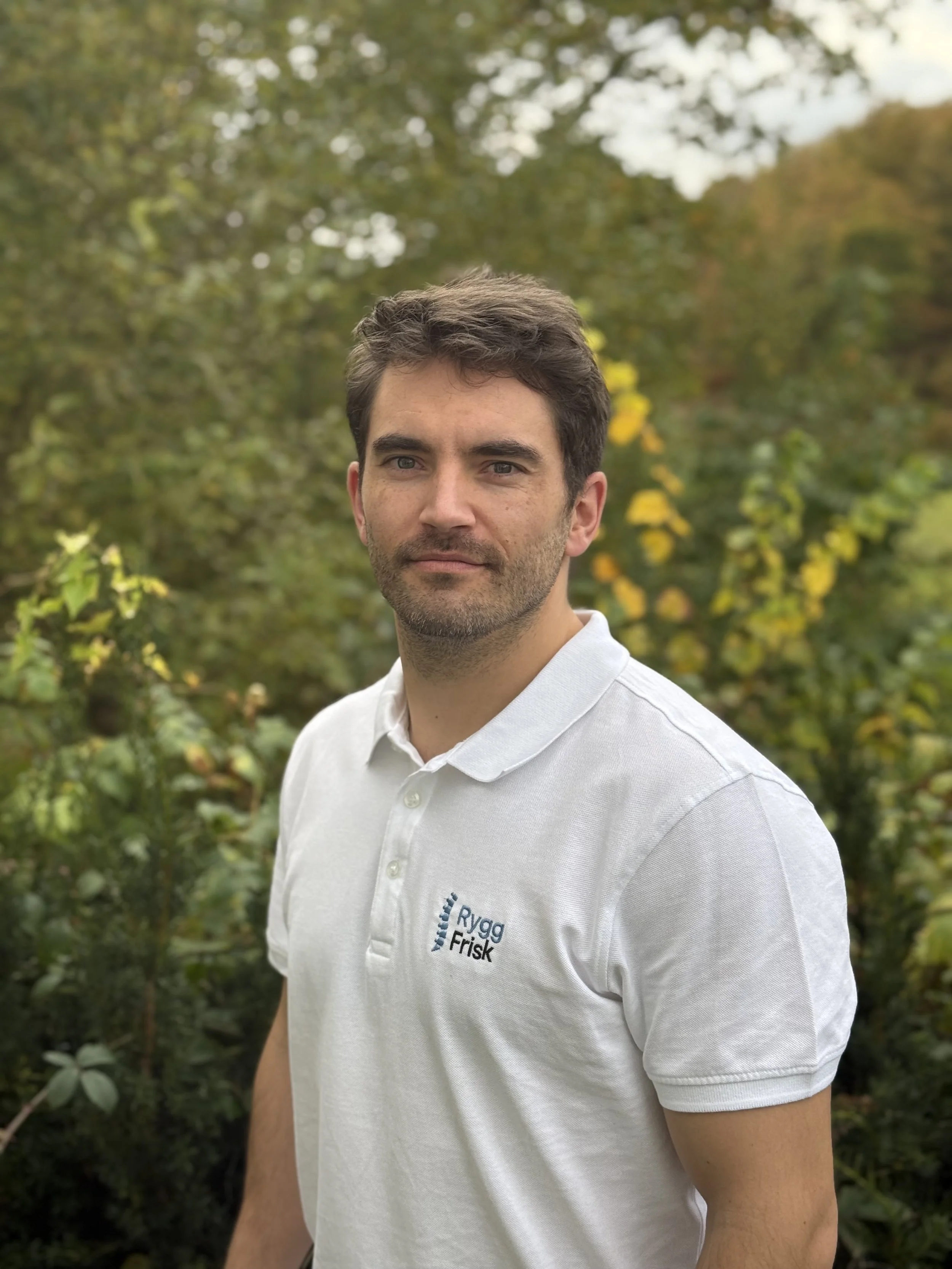 Søramerikansk mann med brunt hår og skjegg, iført hvit polo med logo, står utendørs med grønn vegetasjon i bakgrunnen.