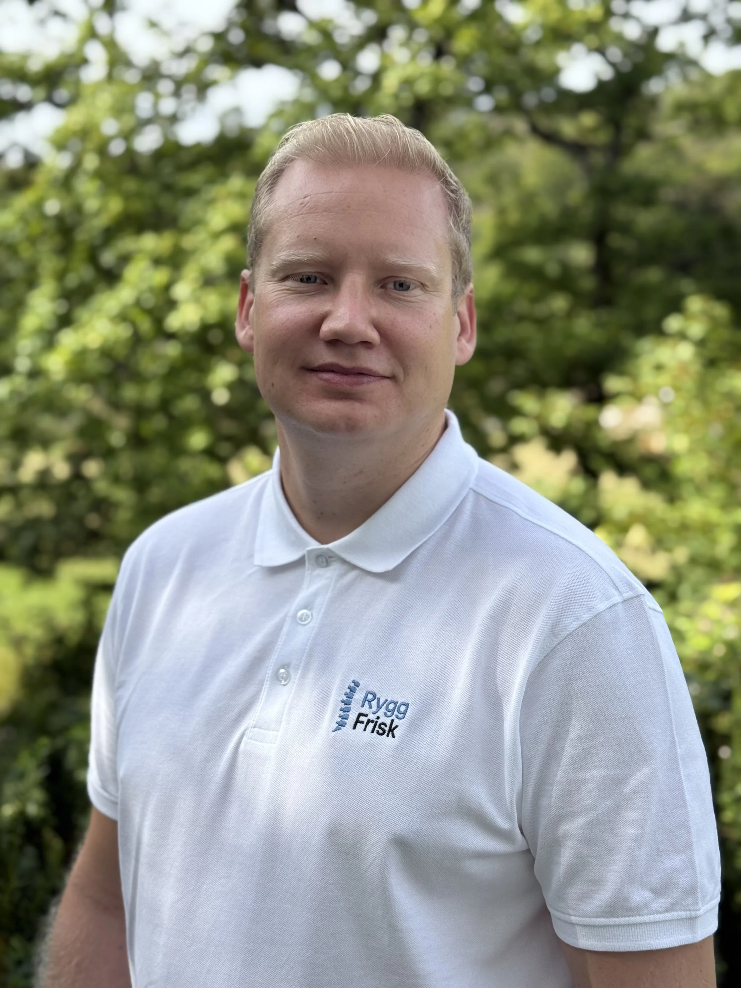 Bildet viser en mann med blond hår iført en hvit polo med logoen 'Rygg Frisk' på, stående utendørs med grønne trær i bakgrunnen.