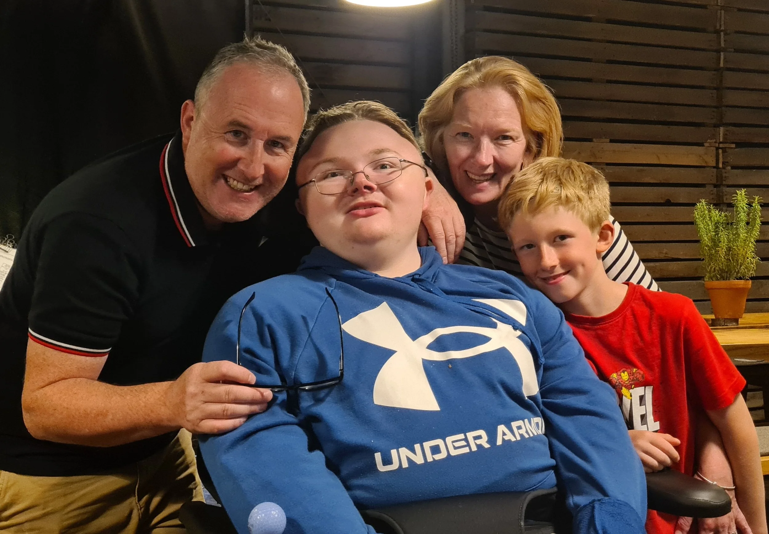 Johanna and Peter with their two boys, Archie is in a wheelchair smiling and Thomas is leaning into Archie. They are indoors, with a wooden wall and a potted plant in the background.