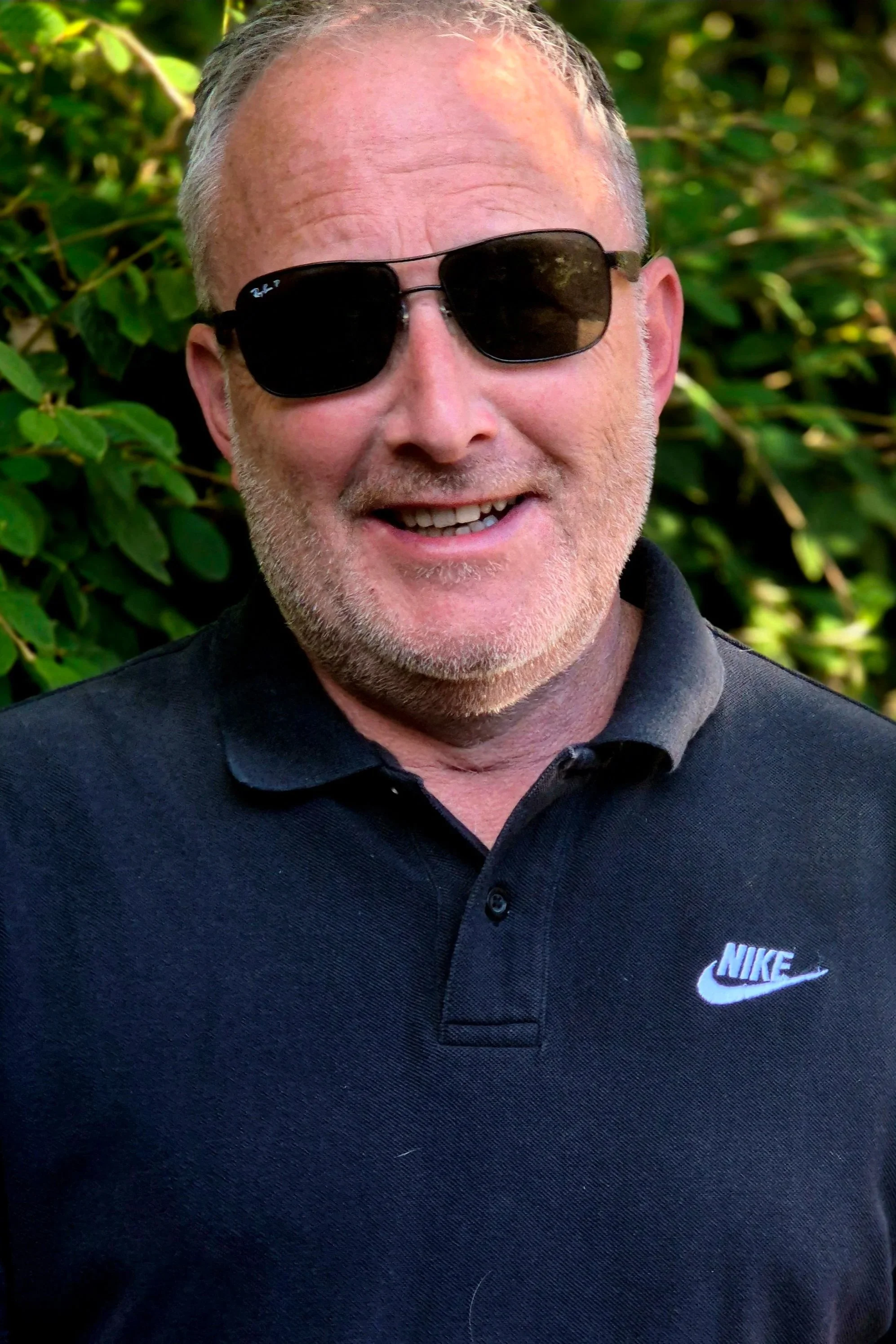 Peter is wearing black sunglasses and a black Nike polo shirt. He is smiling and standing outdoors with green foliage in the background.