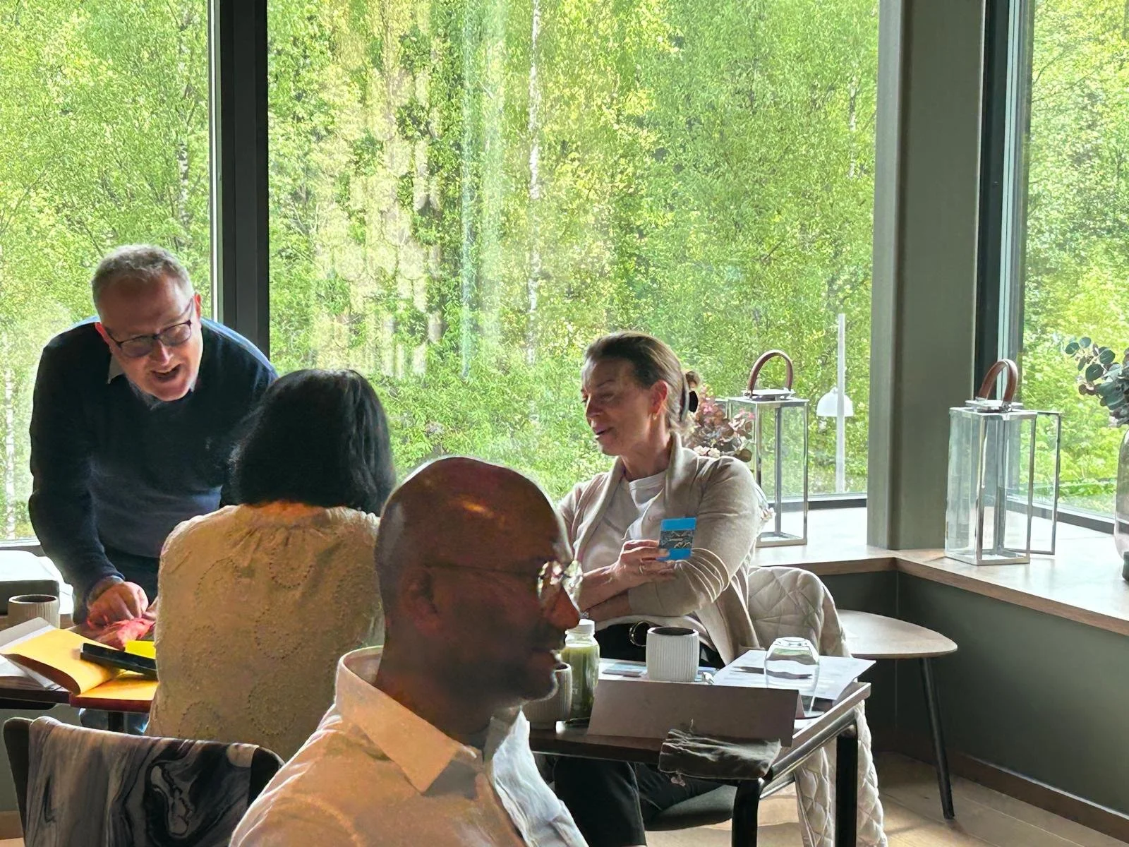 People engaged in a lively conversation in a bright room with large windows overlooking trees, with decorative lanterns on the windowsill.