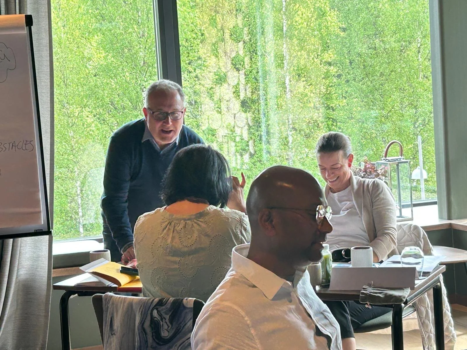 A group of five people sitting around a table, engaging in conversation and laughing, in a room with large windows and greenery outside.