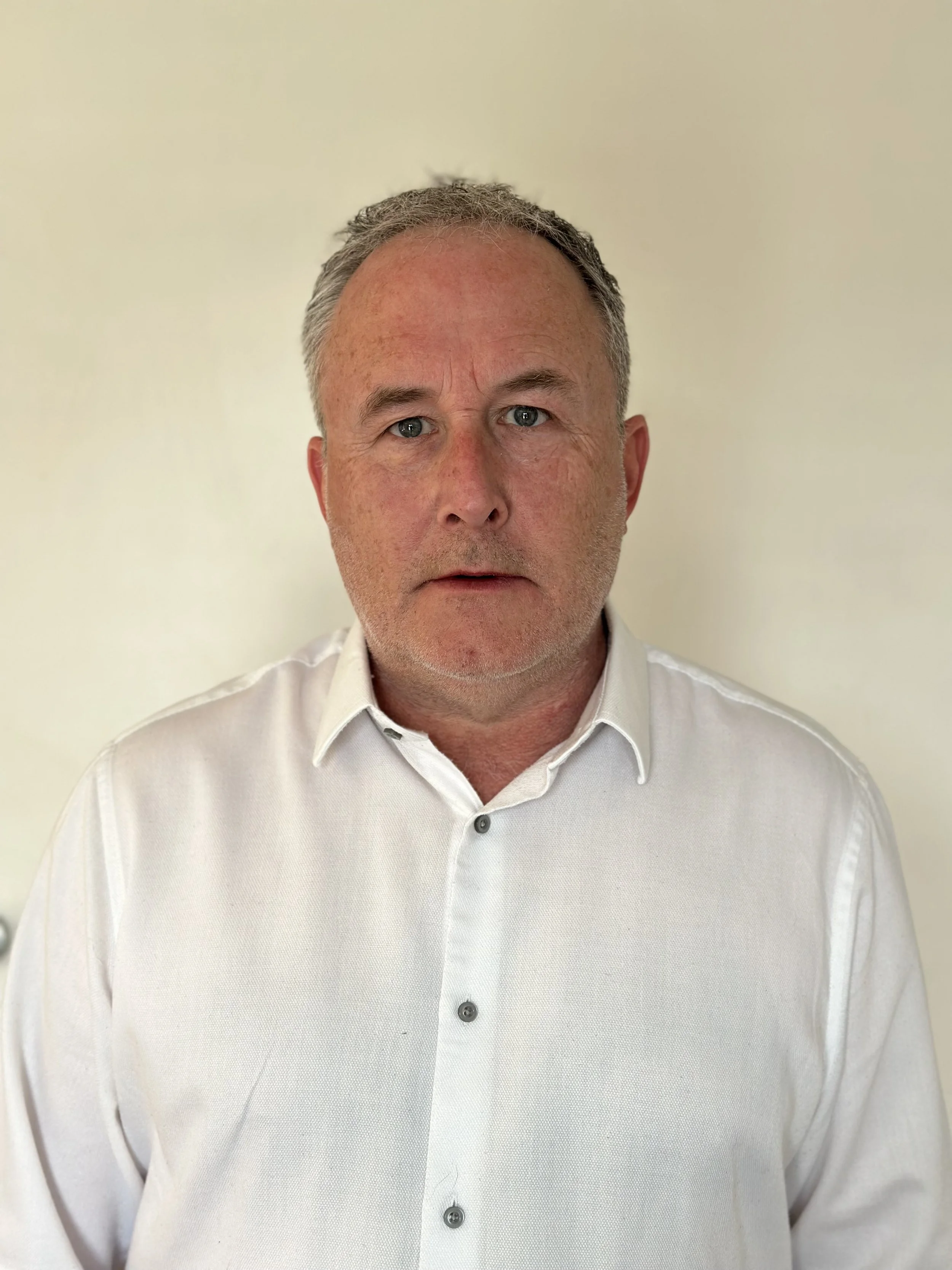 A headshot of Peter, he has  short gray hair and blue eyes wearing a white button-up shirt standing against a plain Offset background.