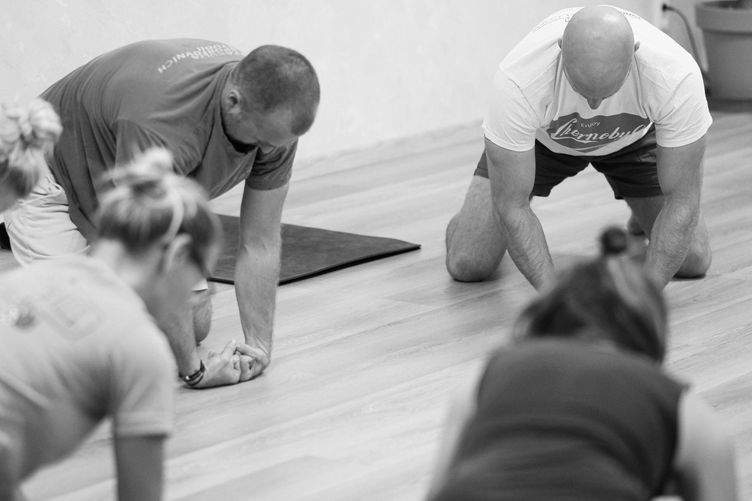 Two men and children engaged in a community yoga or stretching class indoors, with the men demonstrating a stretch or yoga pose on the wooden floor.