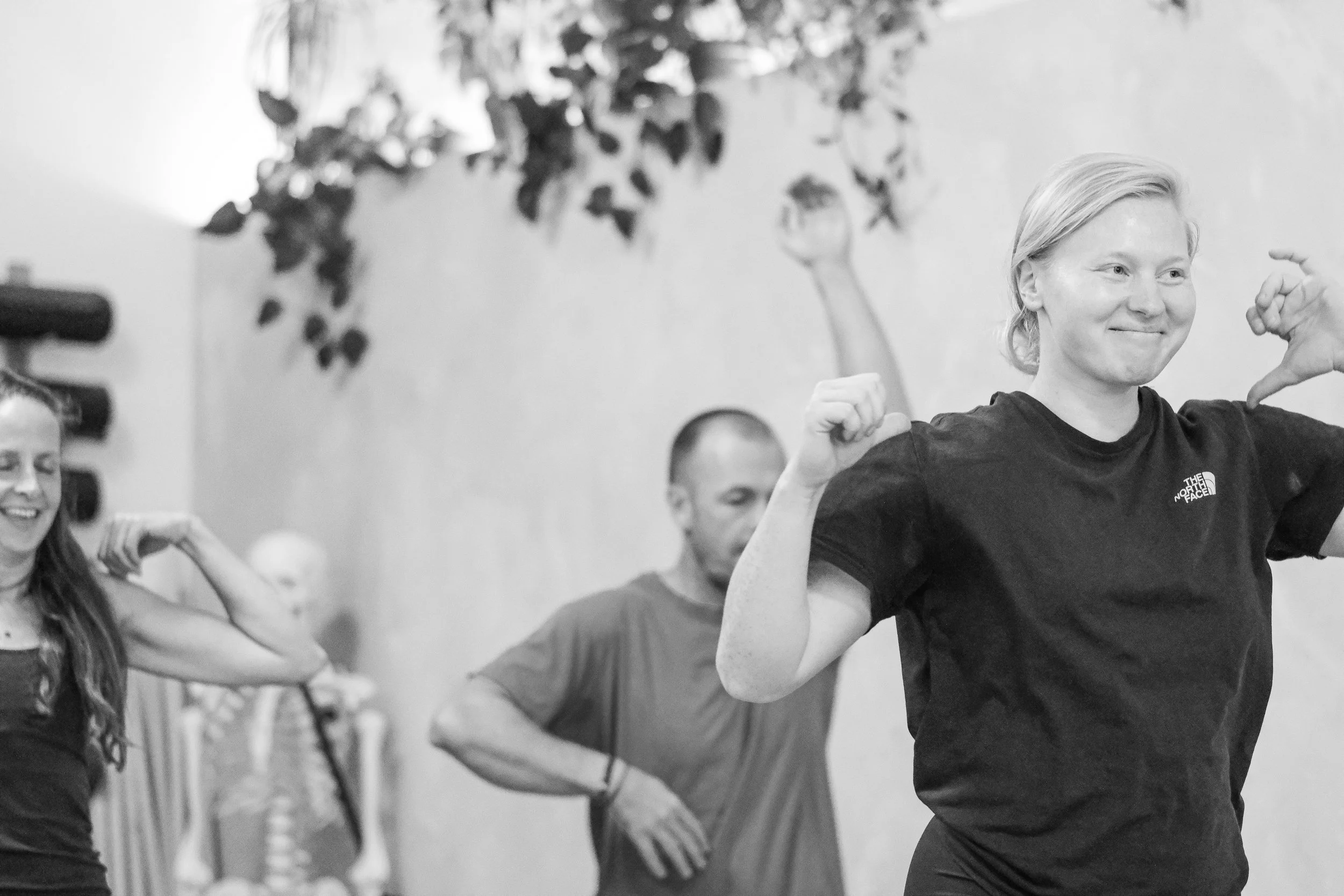 A group of people participating in a fitness or dance class, with a woman in the foreground smiling and making a fist pump gesture with her right hand, wearing a North Face t-shirt. Other participants are blurred in the background, matching her energy.