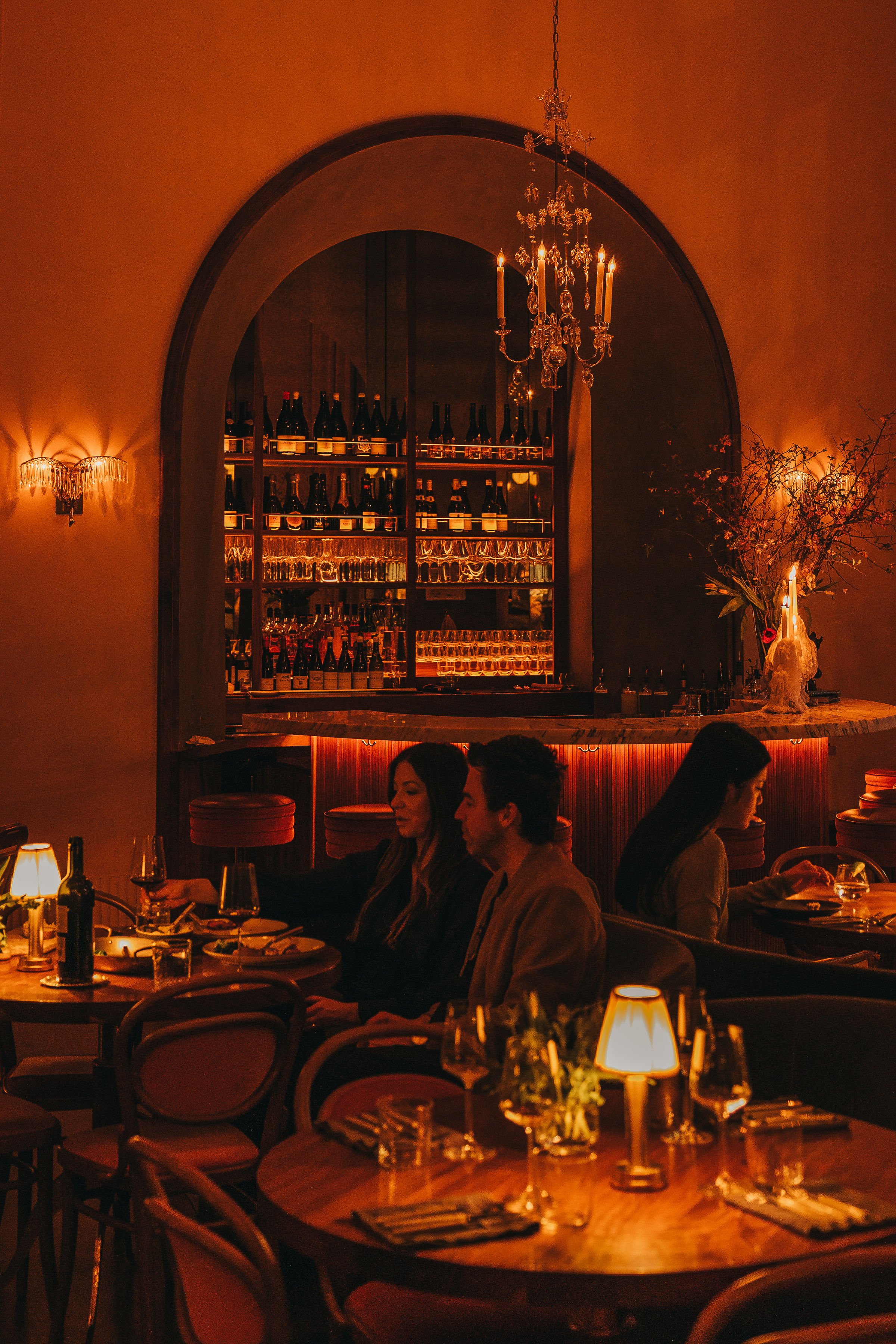 A dimly lit restaurant with a bar area in the background, featuring bottles on shelves, elegant chandeliers, and warm lighting. Couples are dining and conversing at tables.