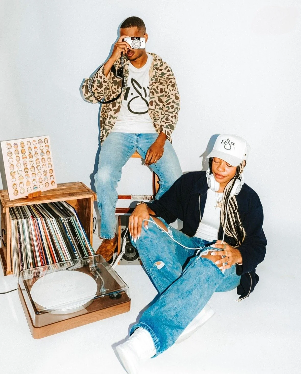 Two young adults enjoying music, one sitting on a chair with a camera, the other sitting on the floor with headphones, surrounded by vinyl records and a turntable.