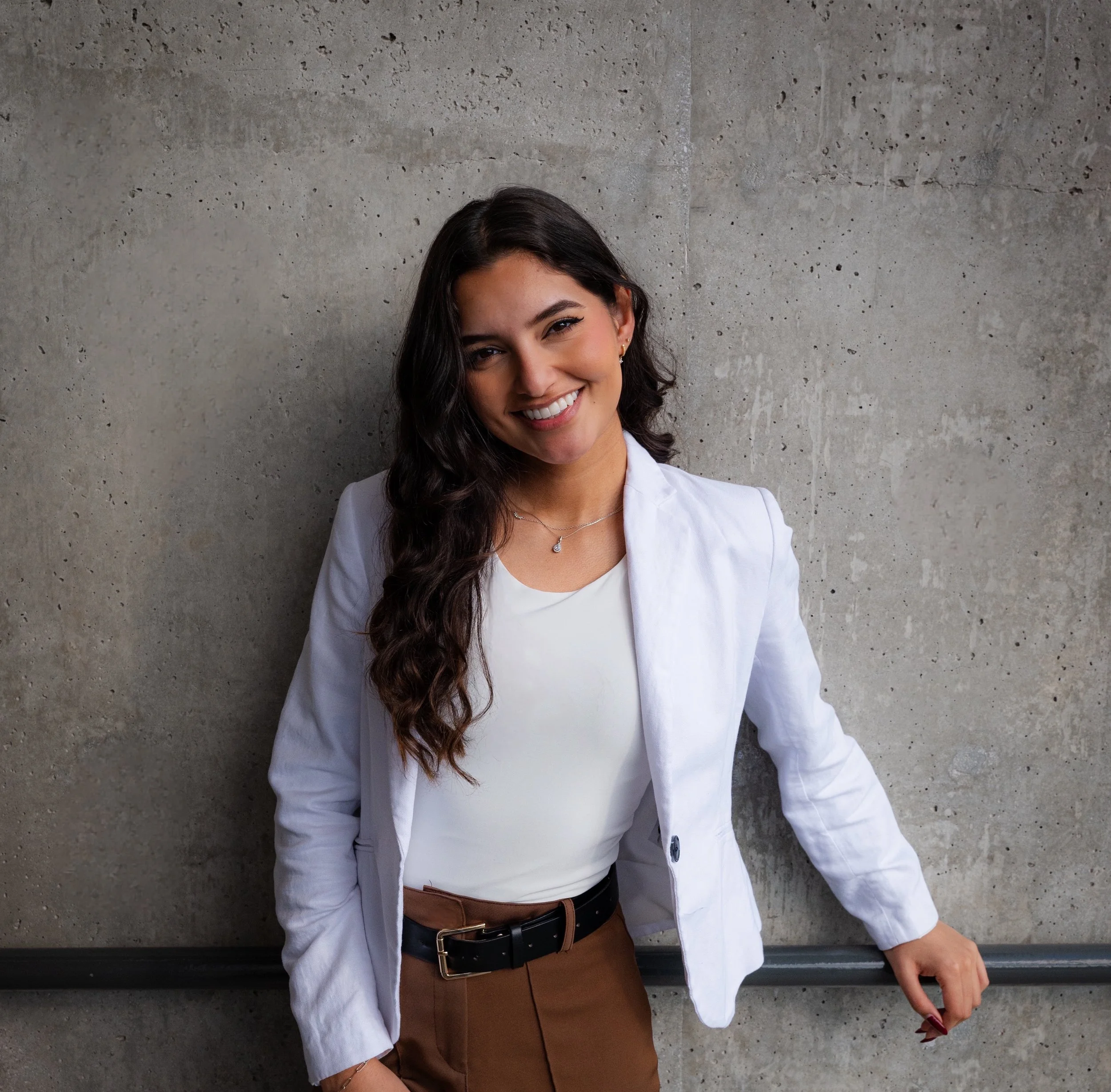 A woman with dark, wavy hair smiling, dressed in a white blazer, brown pants, and a black belt, standing against a concrete wall.