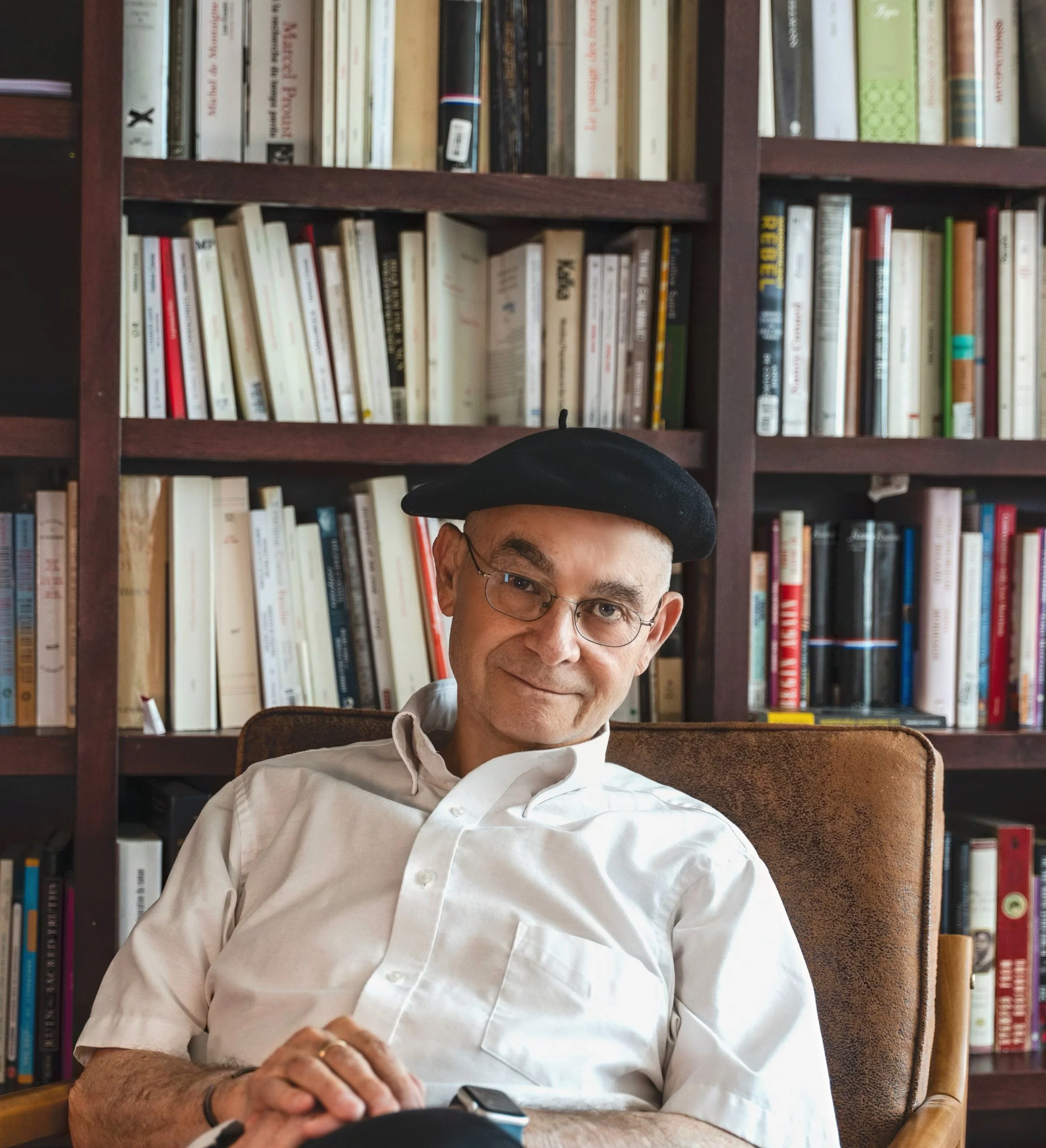 A middle-aged man with glasses and a white shirt sitting in a chair in front of a wooden bookshelf filled with books, wearing a black hat.