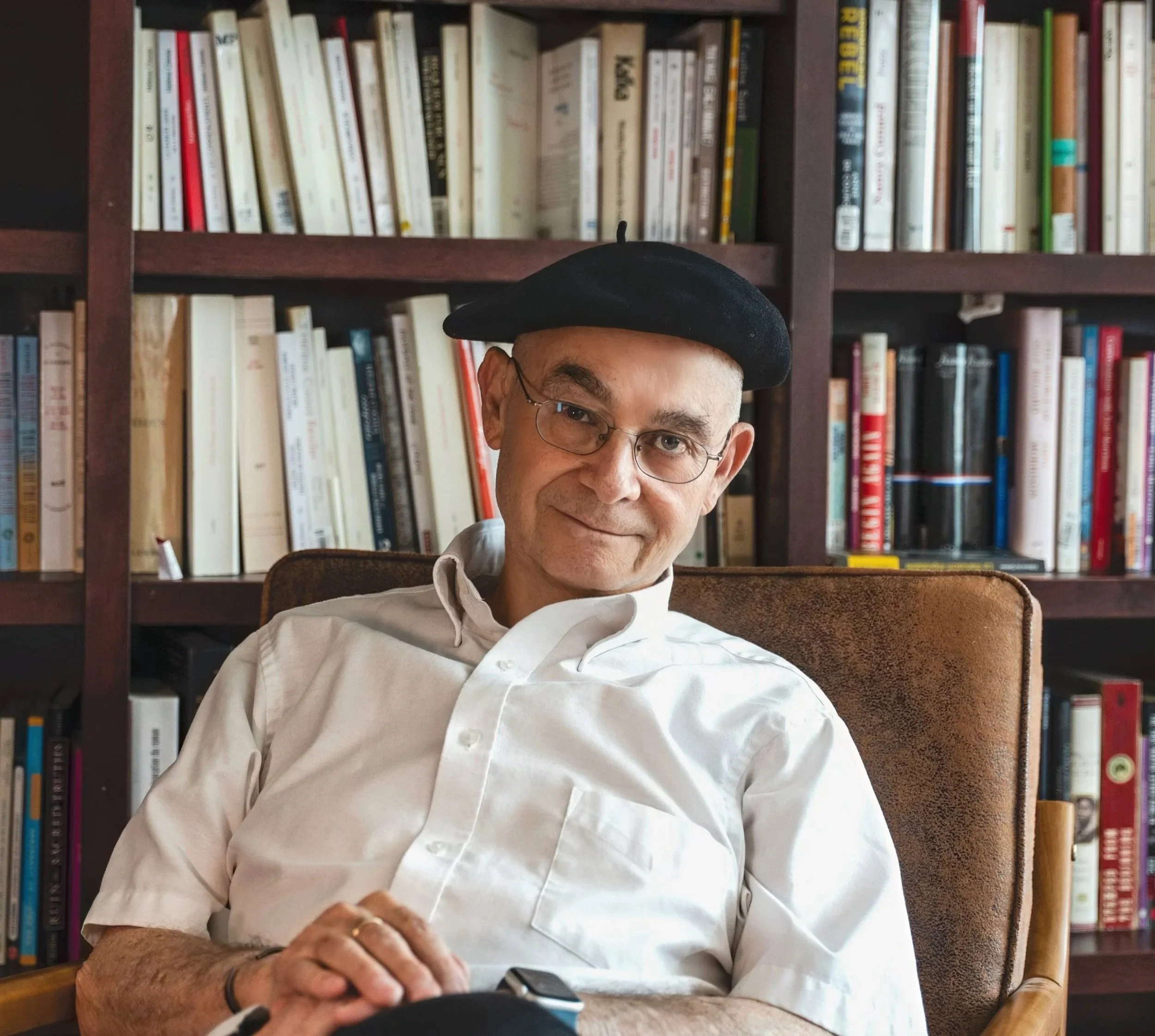 A middle-aged man wearing glasses, a black beret, and a white button-up shirt sitting in front of a bookshelf filled with various books. Part of his face and hand are visible.