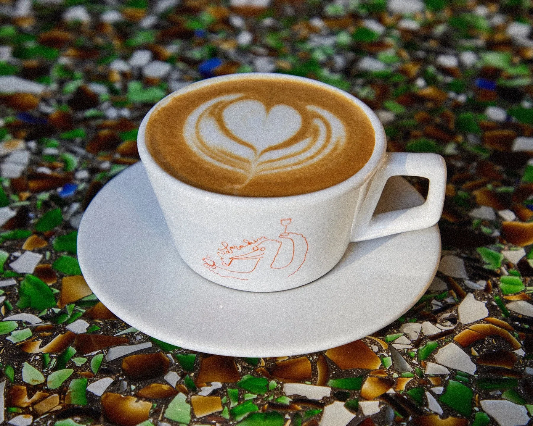 A cup of coffee with latte art featuring a heart shape, placed on a matching white saucer with a minimalist line drawing of a person. The background is composed of colorful mosaic tiles.